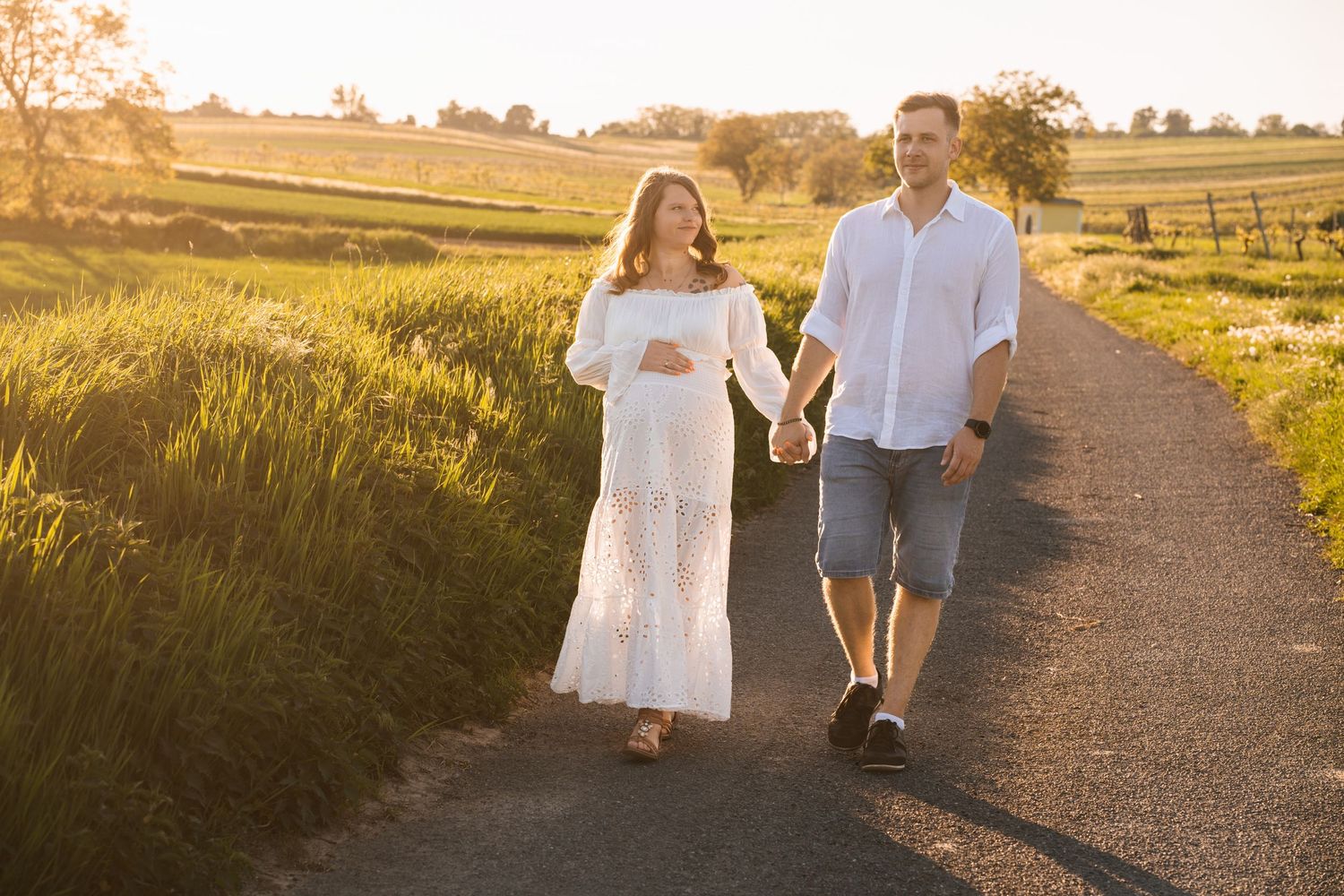 Couple walking hand in hand on a dirt path through golden wheat fields at sunset.