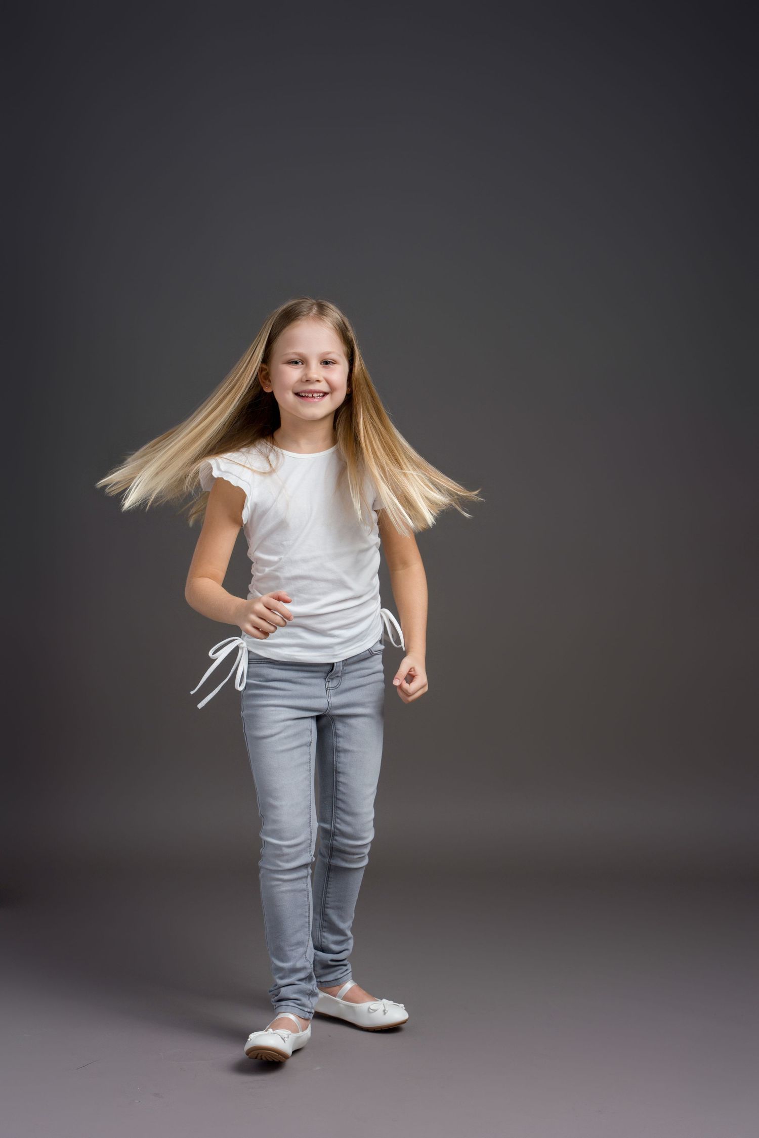 Child with flowing blonde hair poses energetically in white top and jeans against grey background.