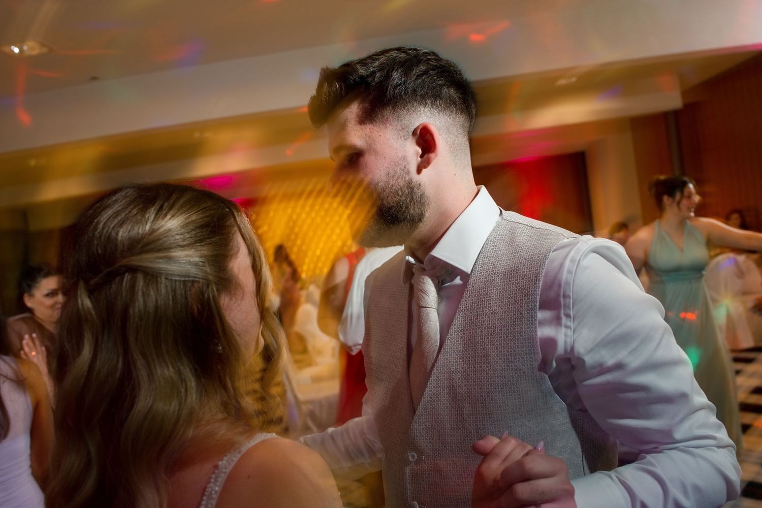 Couple sharing an intimate dance at a wedding reception with colorful lights and guests in the background.