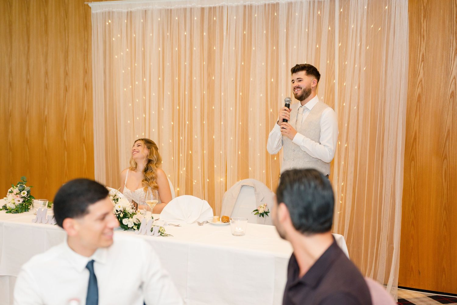 Man giving a speech at a wedding reception with bride seated at the head table, elegant floral decor.