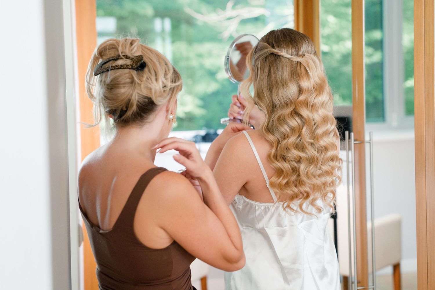 Bridesmaid in brown dress helping blonde bride in white gown prepare, viewed from behind near mirror.