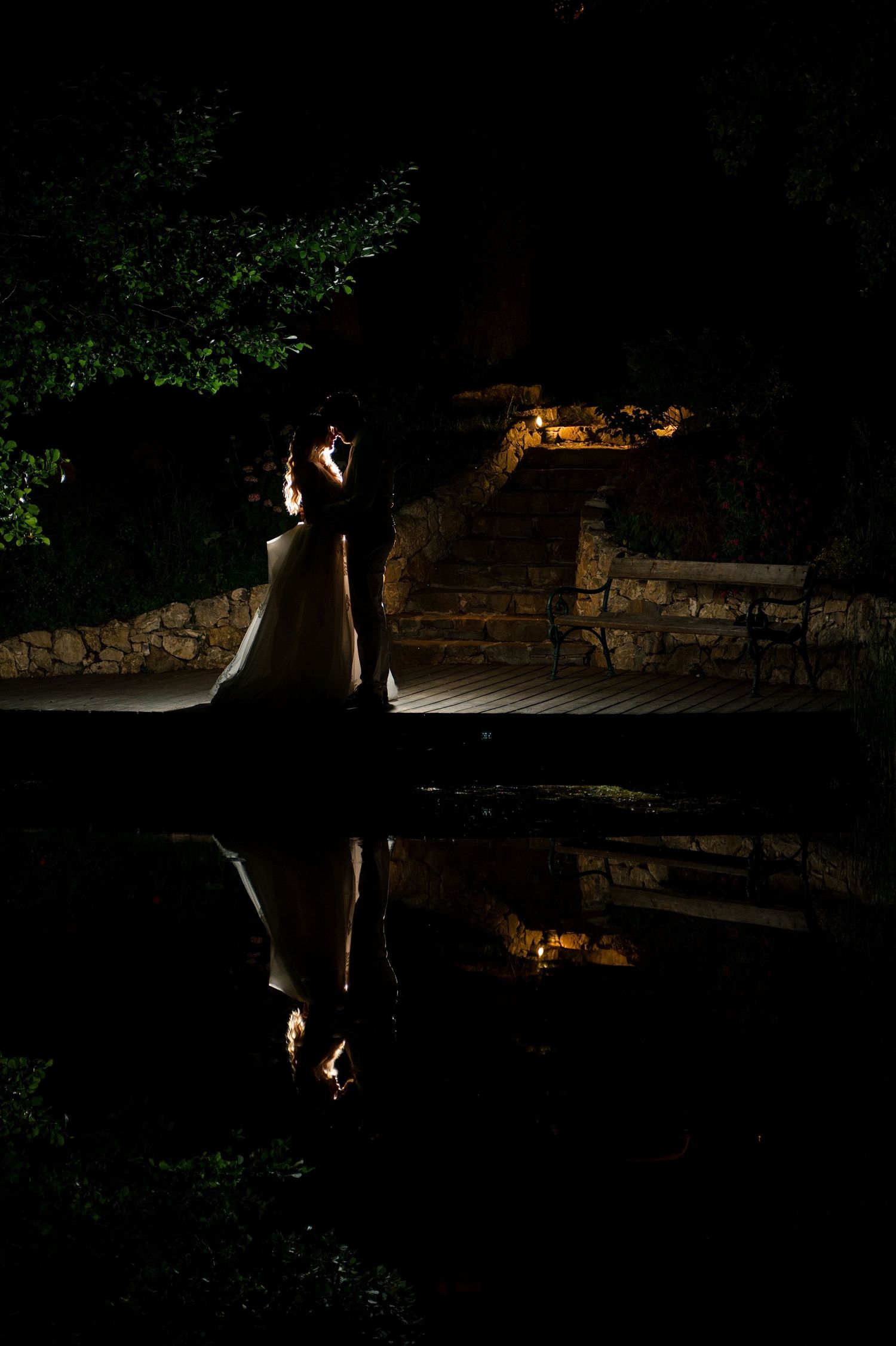 Bride and groom silhouette reflected in a pool at night, surrounded by stone steps and lush greenery.