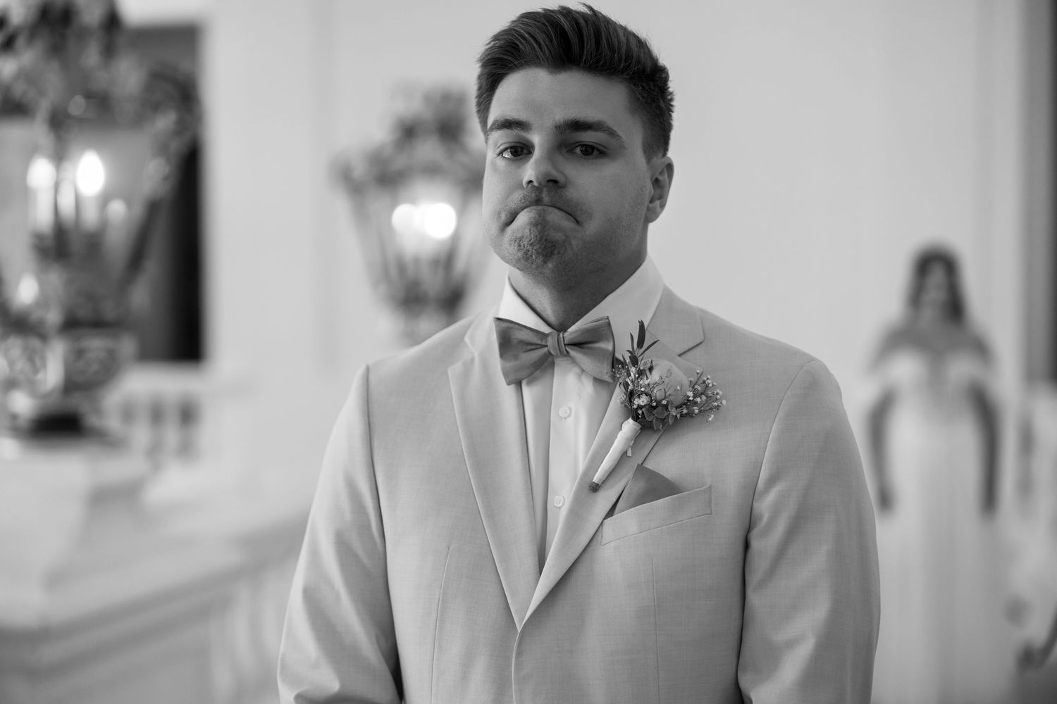 Groom in light colored suit with bow tie at indoor venue.