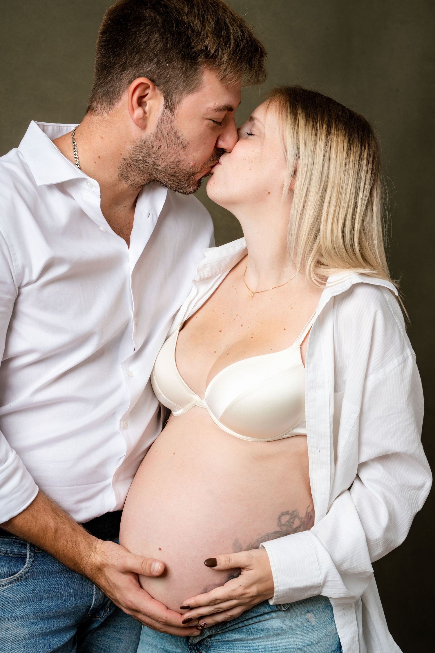 Intimate maternity photoshoot featuring couple in white shirts and jeans against moody backdrop.