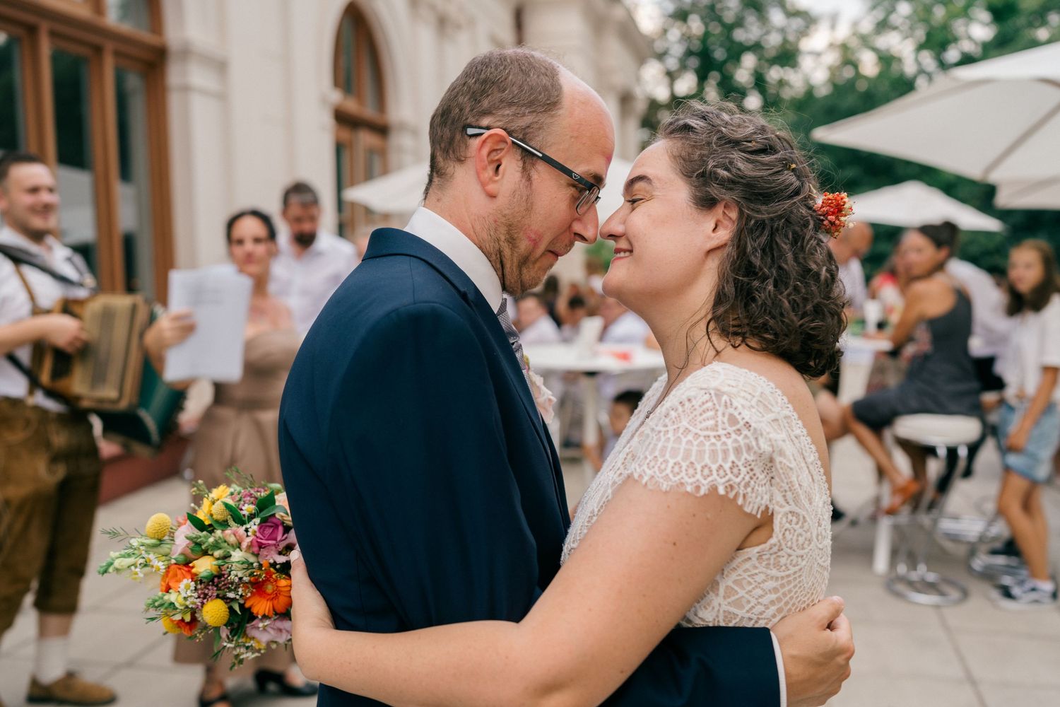 A couple shares a romantic moment at an outdoor wedding, the groom holding a colorful bouquet while guests celebrate in the background.