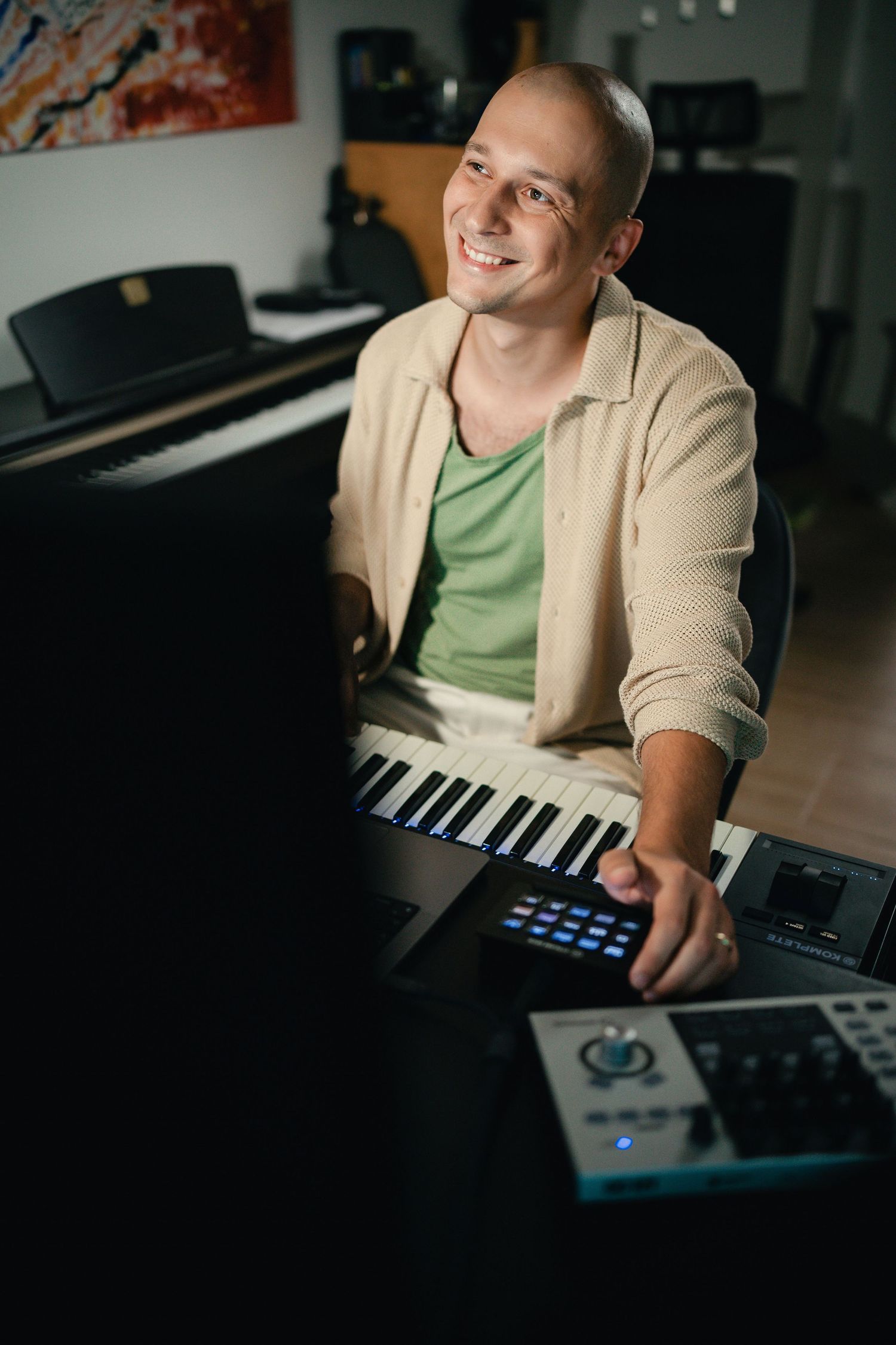 A musician in a beige sweater and green shirt sits at a digital keyboard controller in a dimly lit studio.