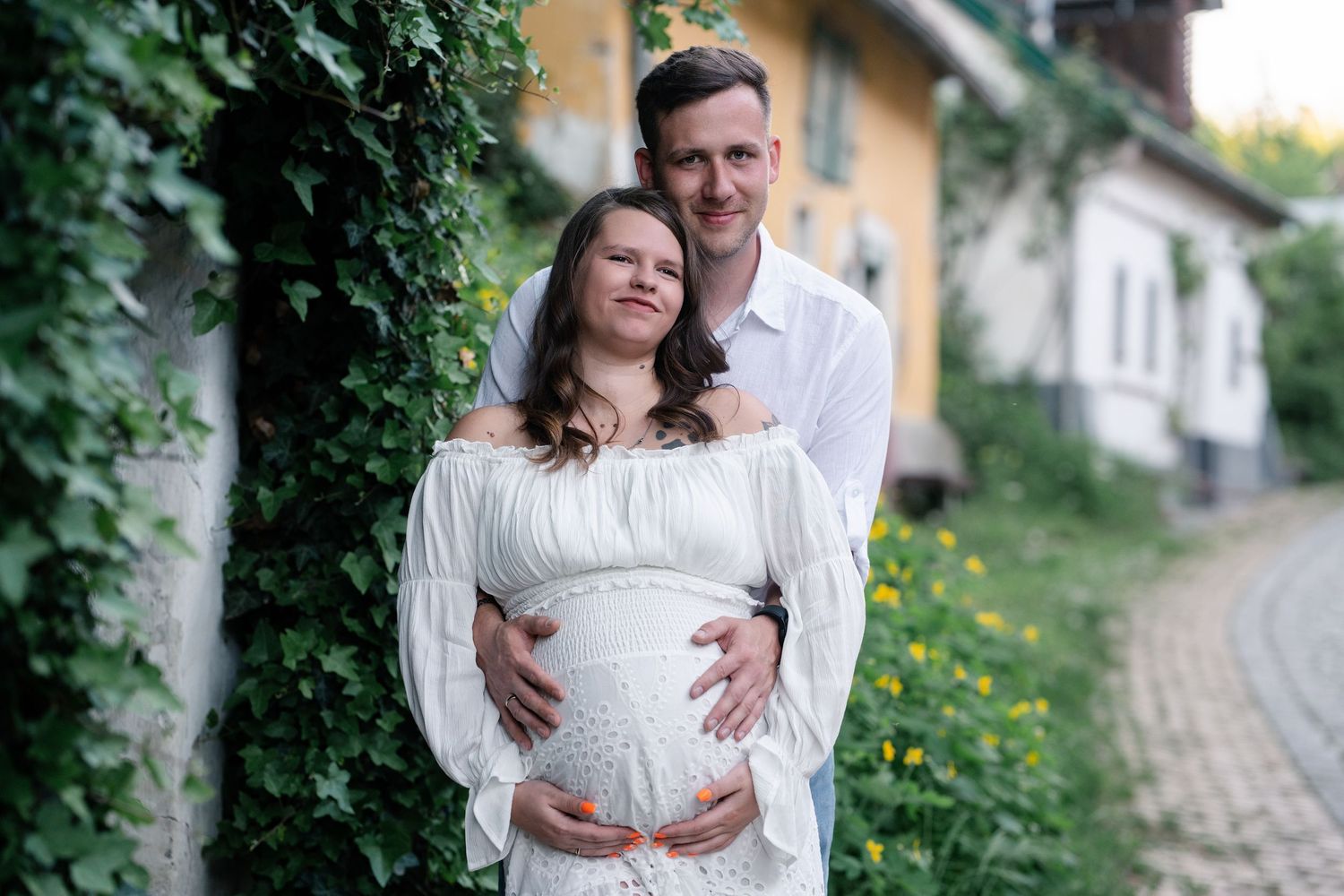 A couple in white embraces during a romantic outdoor maternity photo session with greenery and stone path backdrop.
