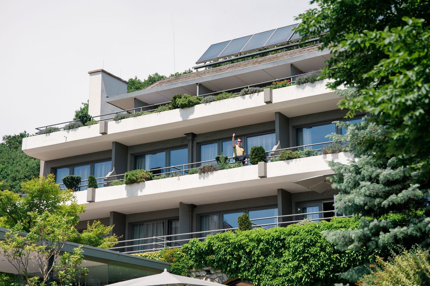 Modern multi-story apartment building with white balconies, lush greenery, and rooftop solar panels on a sunny day.