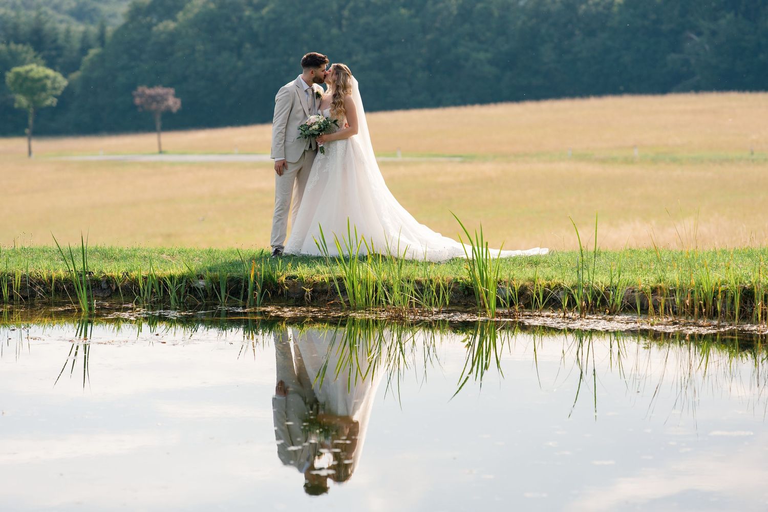 Bride and groom kissing by a reflective pond with lush green fields and trees in the background on their wedding day.