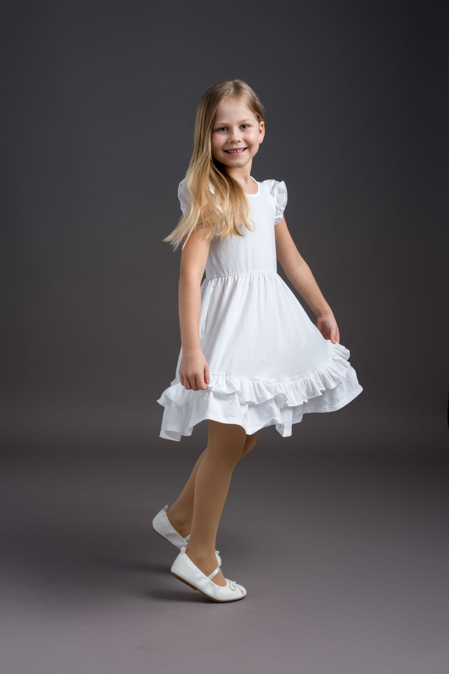 Child wearing white dress and brown tights twirls playfully in a studio setting.