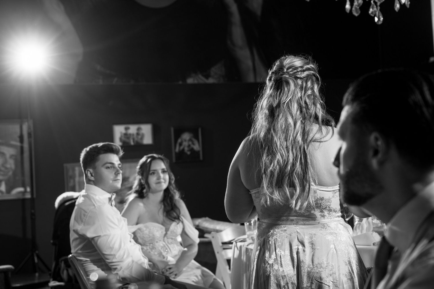 Black and white wedding photo of bride and groom seated at reception table with guests nearby.