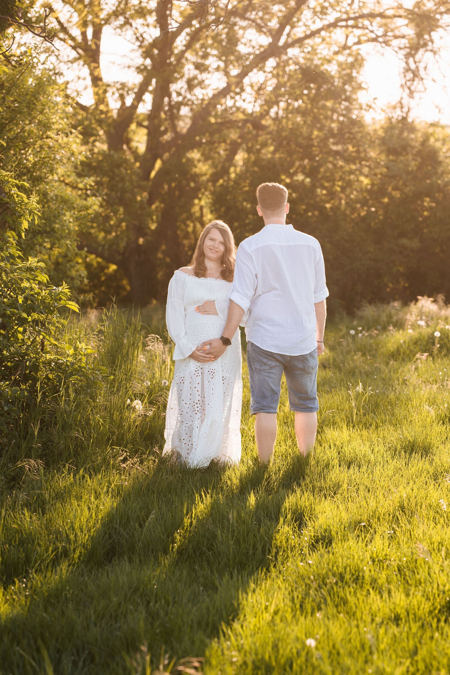 A couple stands together in a sunlit meadow at golden hour casting long shadows on the grass.