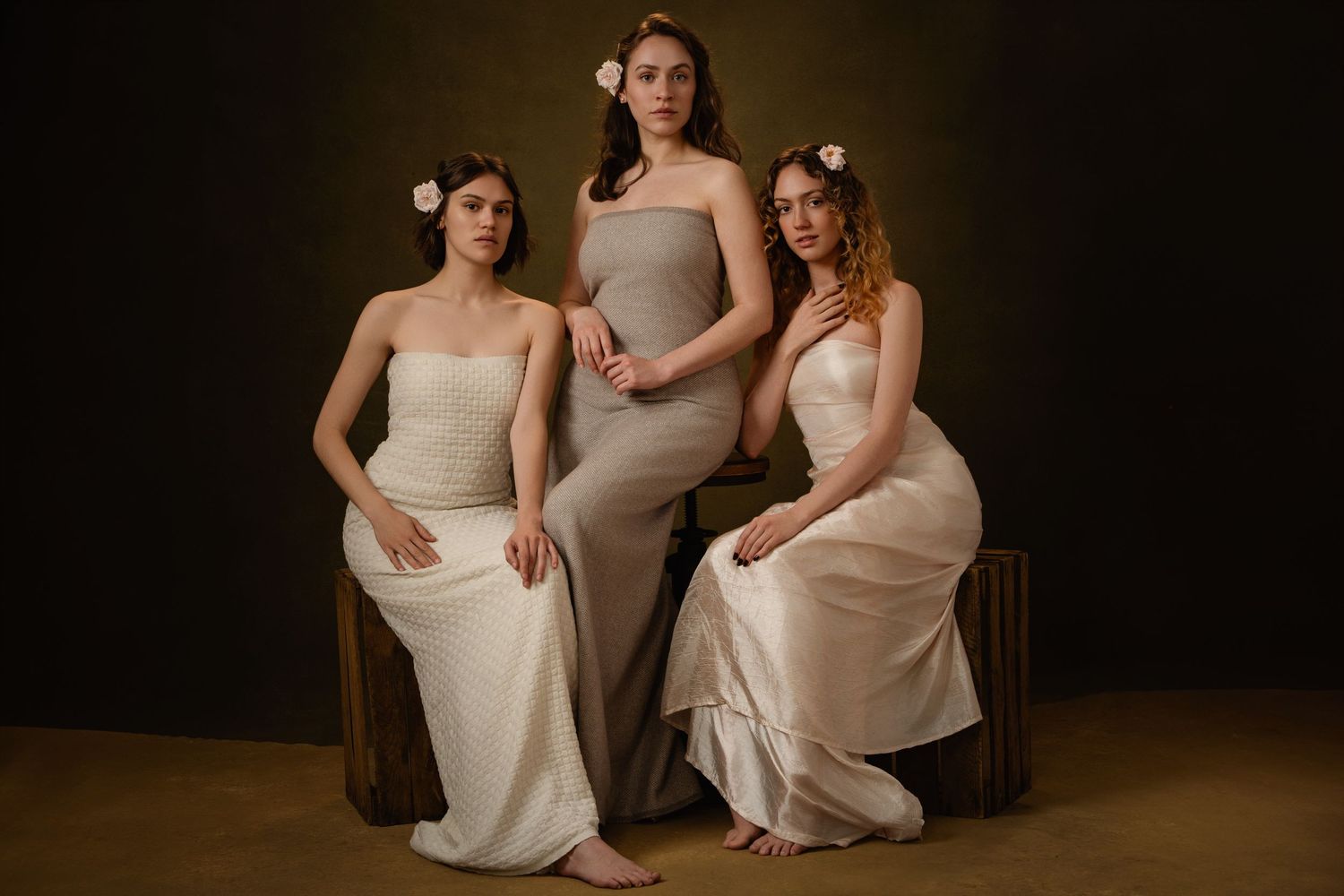 Three people in cream-colored dresses pose together against a dark background in an elegant portrait series.