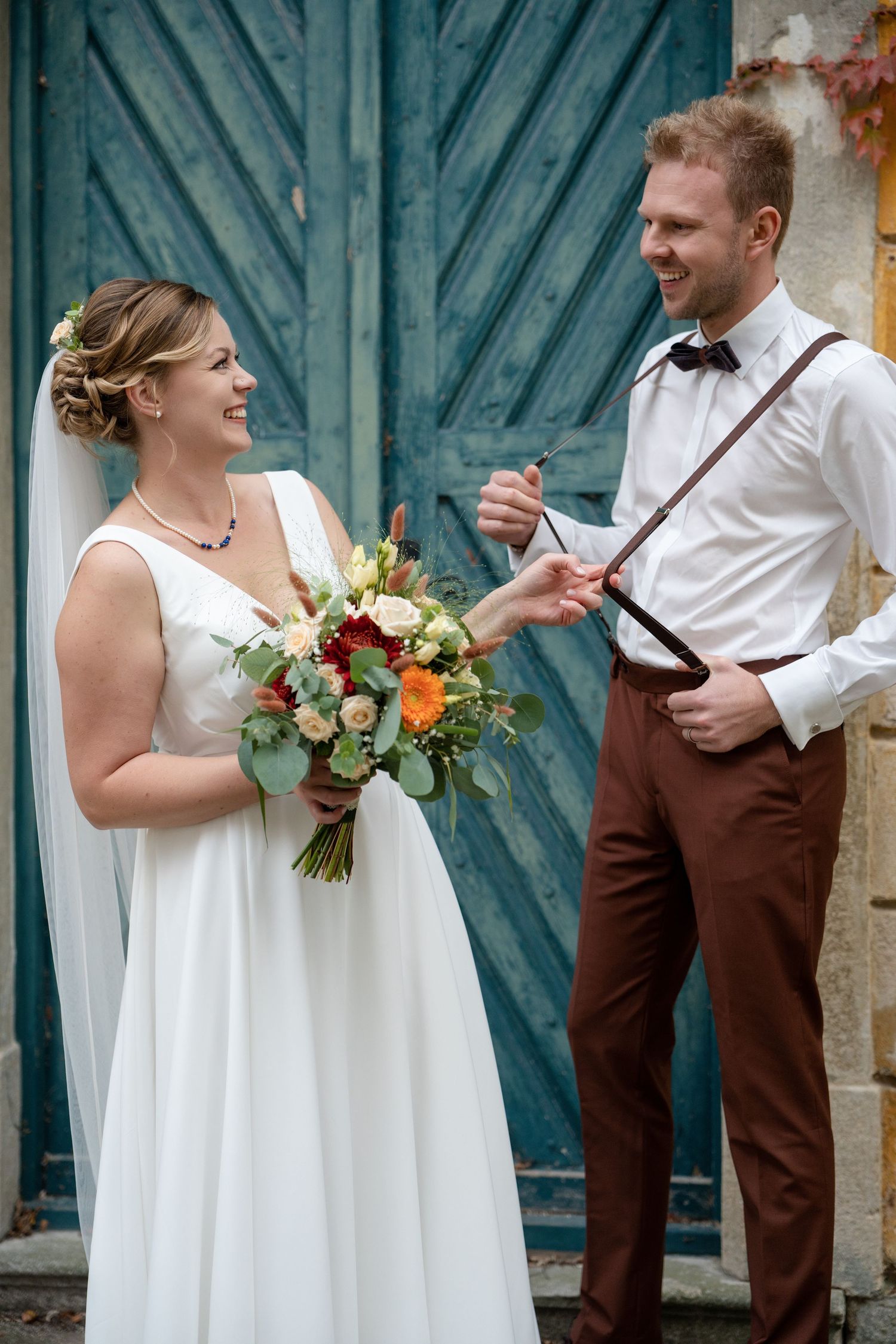 Wedding couple in white dress and brown vest posing against blue wooden doors.