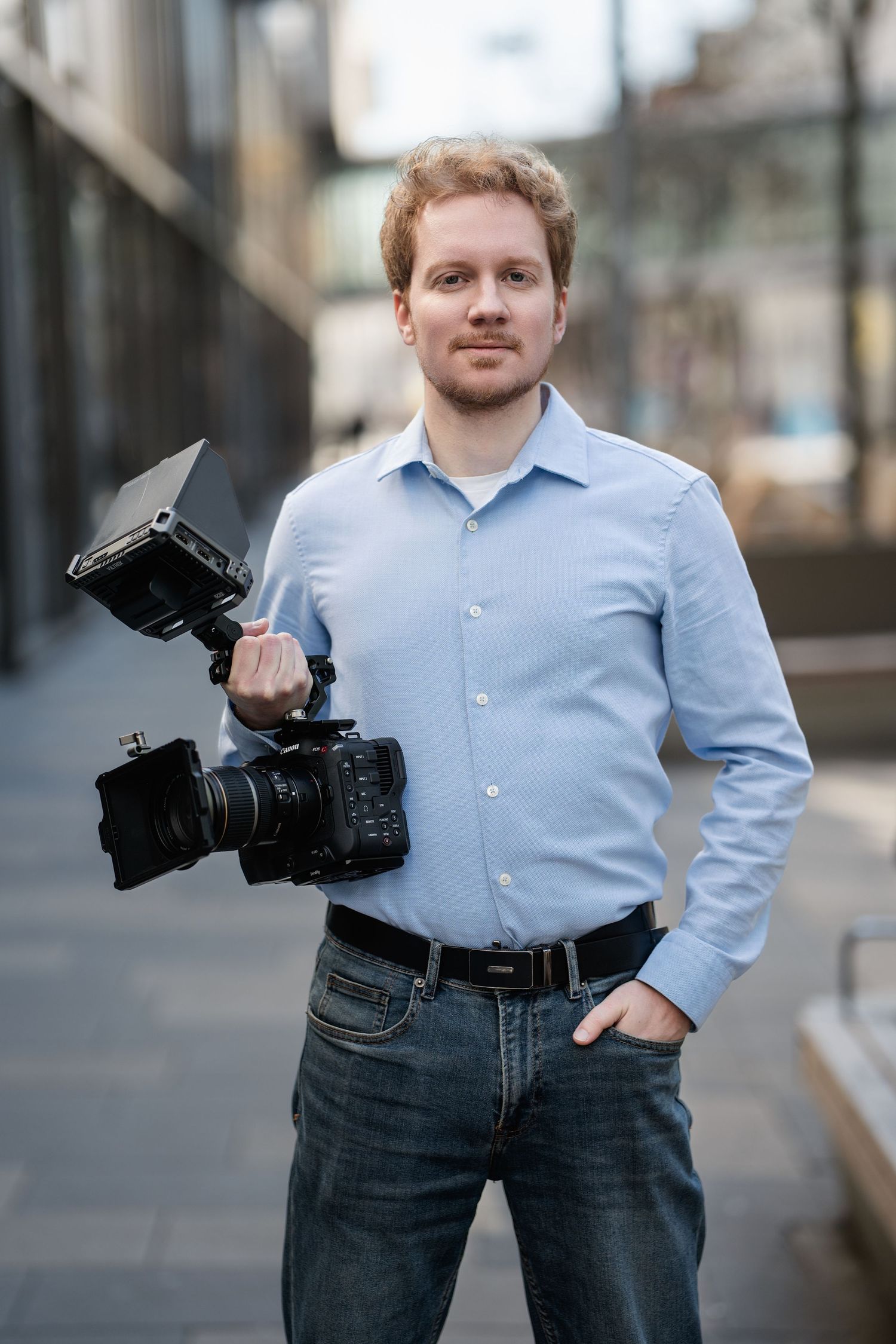Professional videographer in light blue shirt and jeans holding a camera on a city street.