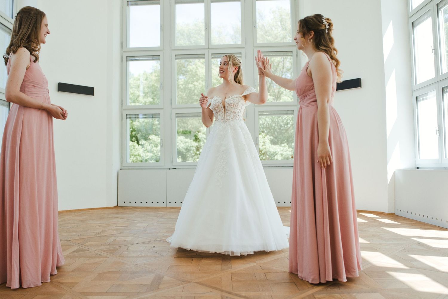 Bride in white gown with two bridesmaids in pink dresses celebrating in a bright room with hardwood floors.