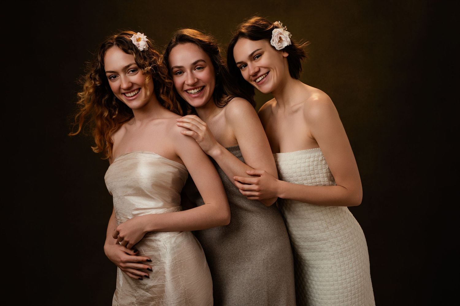 Three people in elegant strapless gowns laugh together against a dark background during a formal photoshoot.