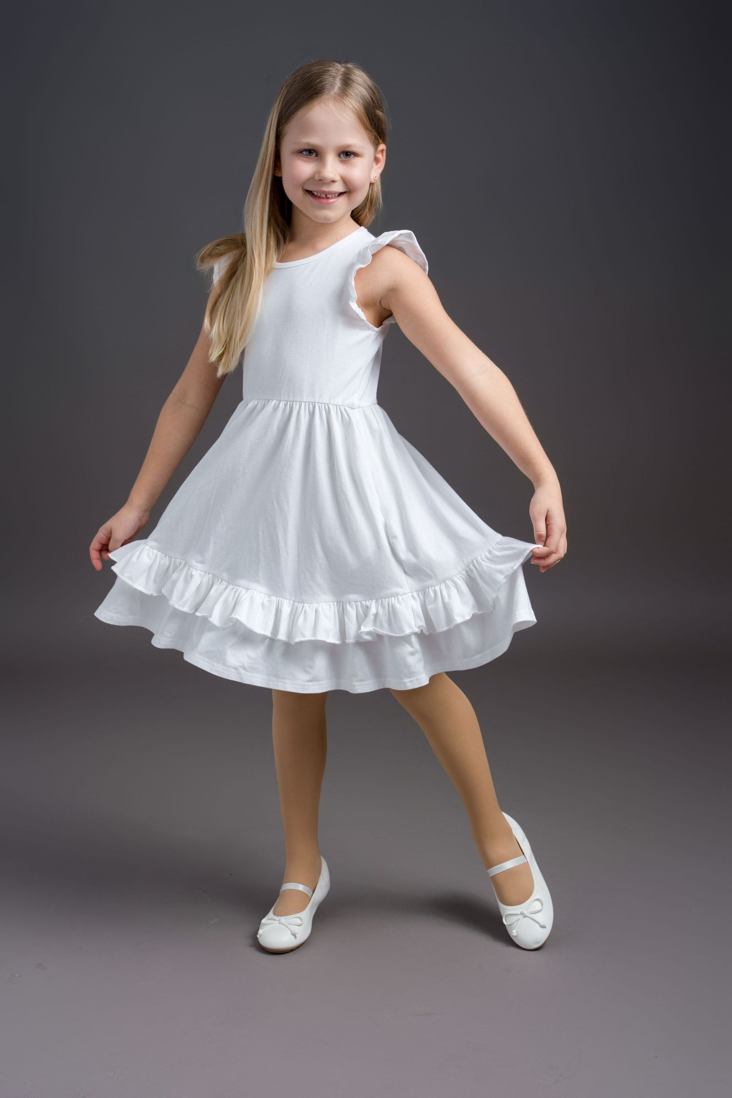 A child in a white ruffled dress twirls and dances in front of a gray studio backdrop.