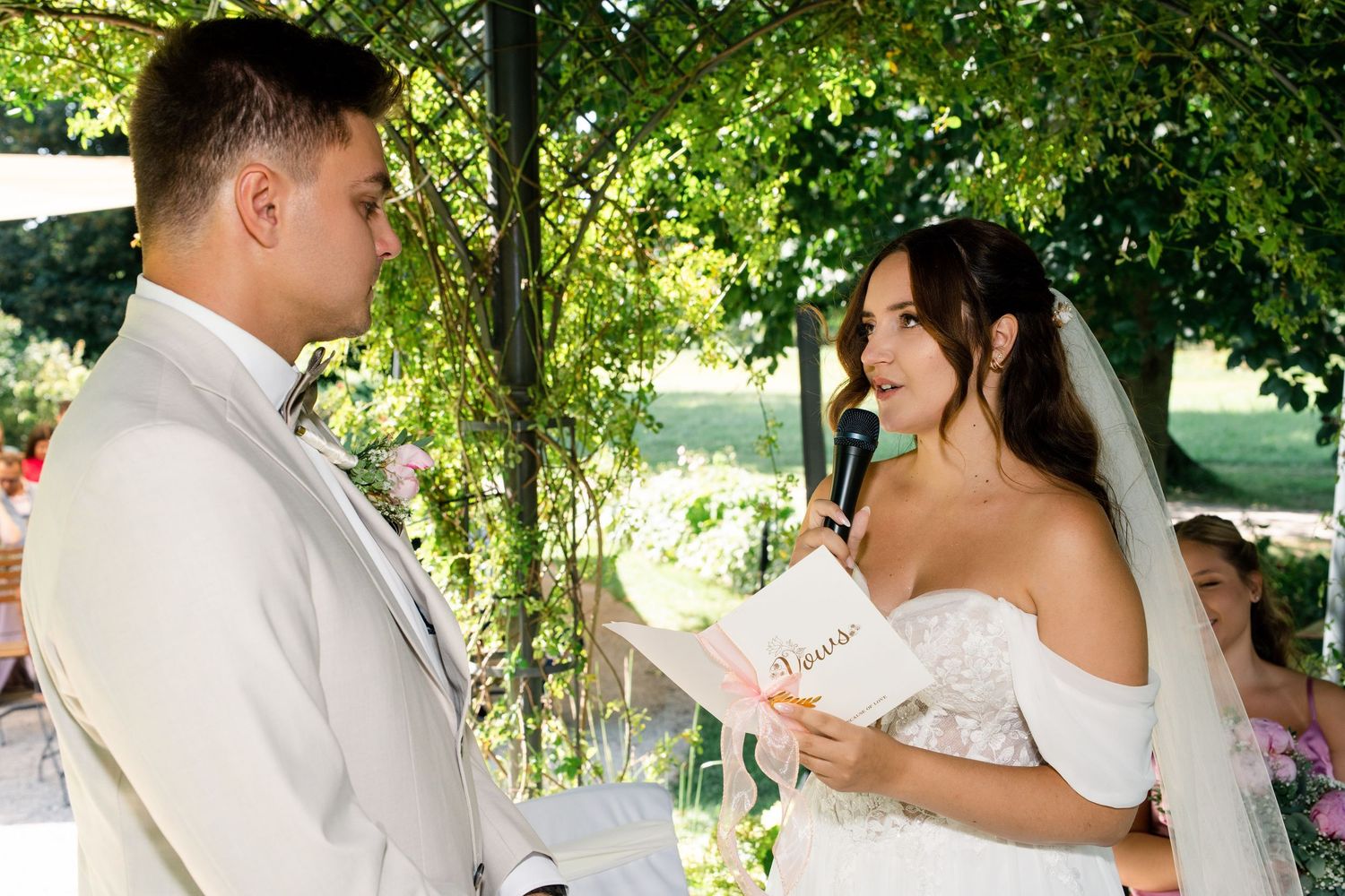 Newlyweds exchange vows under lush green trees during a summer wedding ceremony.
