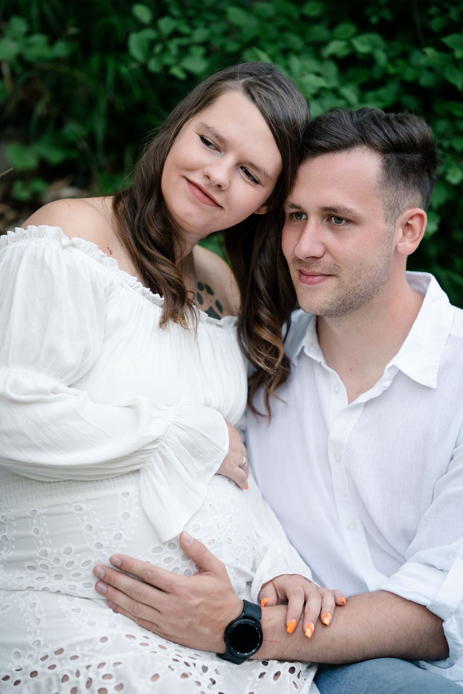 A couple in white clothing poses intimately together during an outdoor photo session with lush greenery in background.