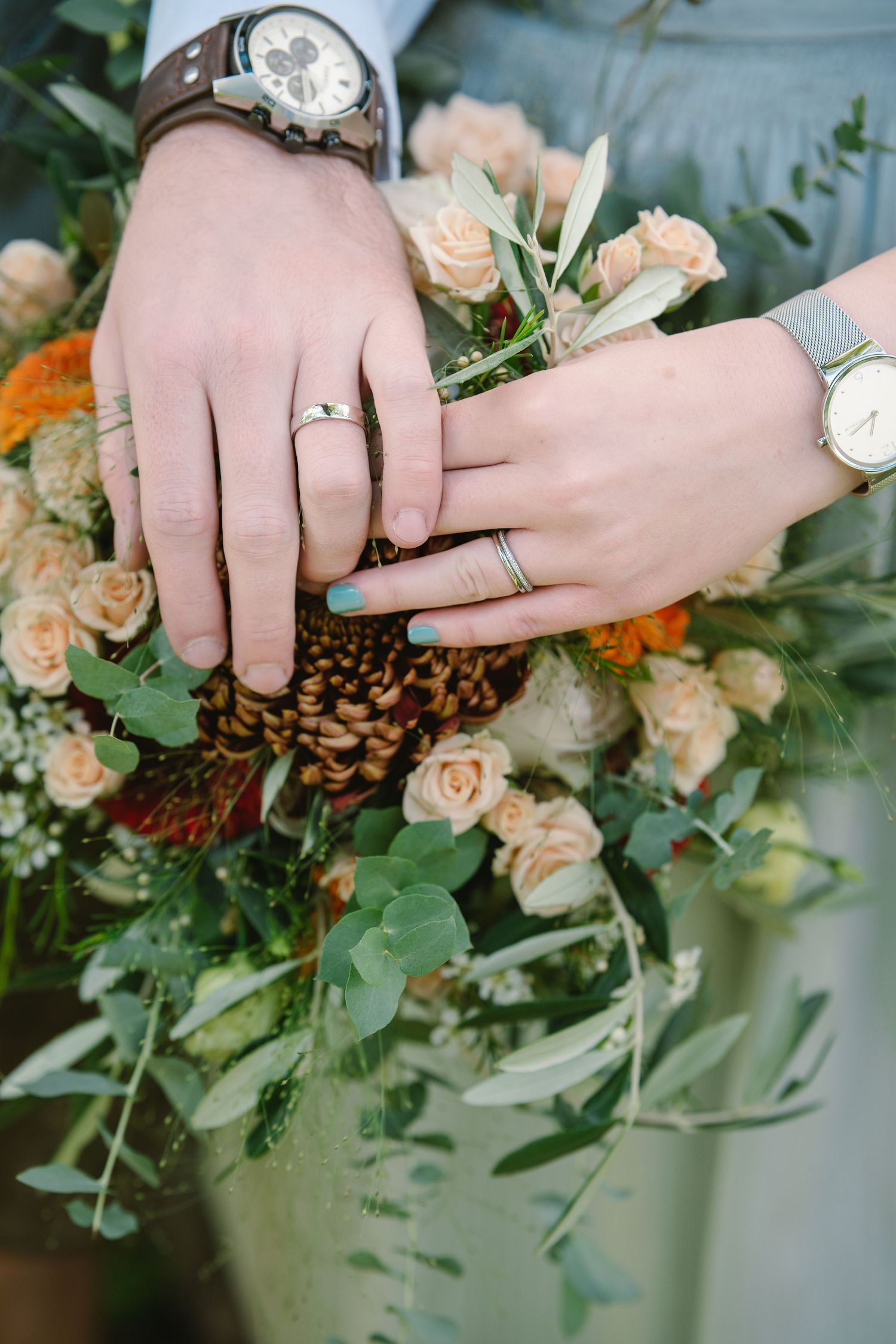 Close-up of hands wearing watches and rings resting on a peach and orange floral bouquet with greenery.
