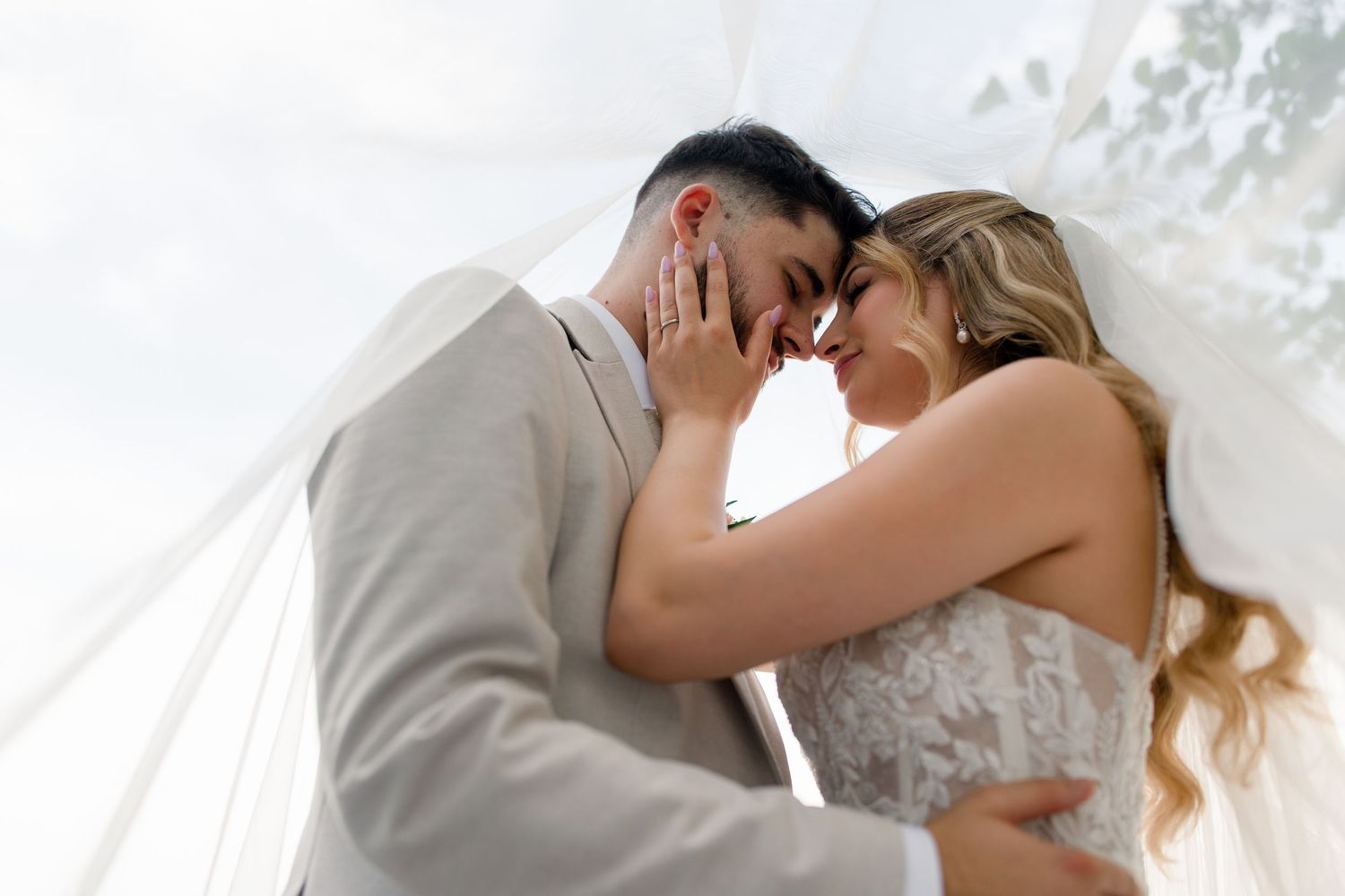 Romantic bride and groom touching foreheads under a white veil on their wedding day.