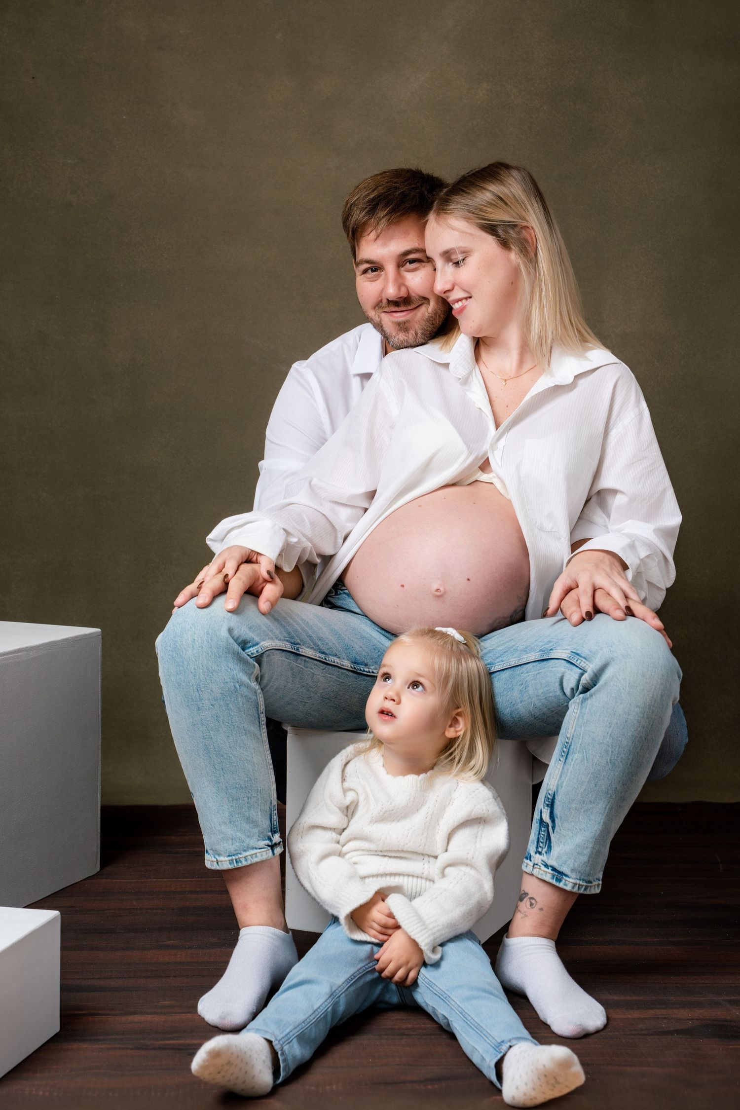 Tender family moments captured in a studio setting with casual denim and white clothing against dark background.