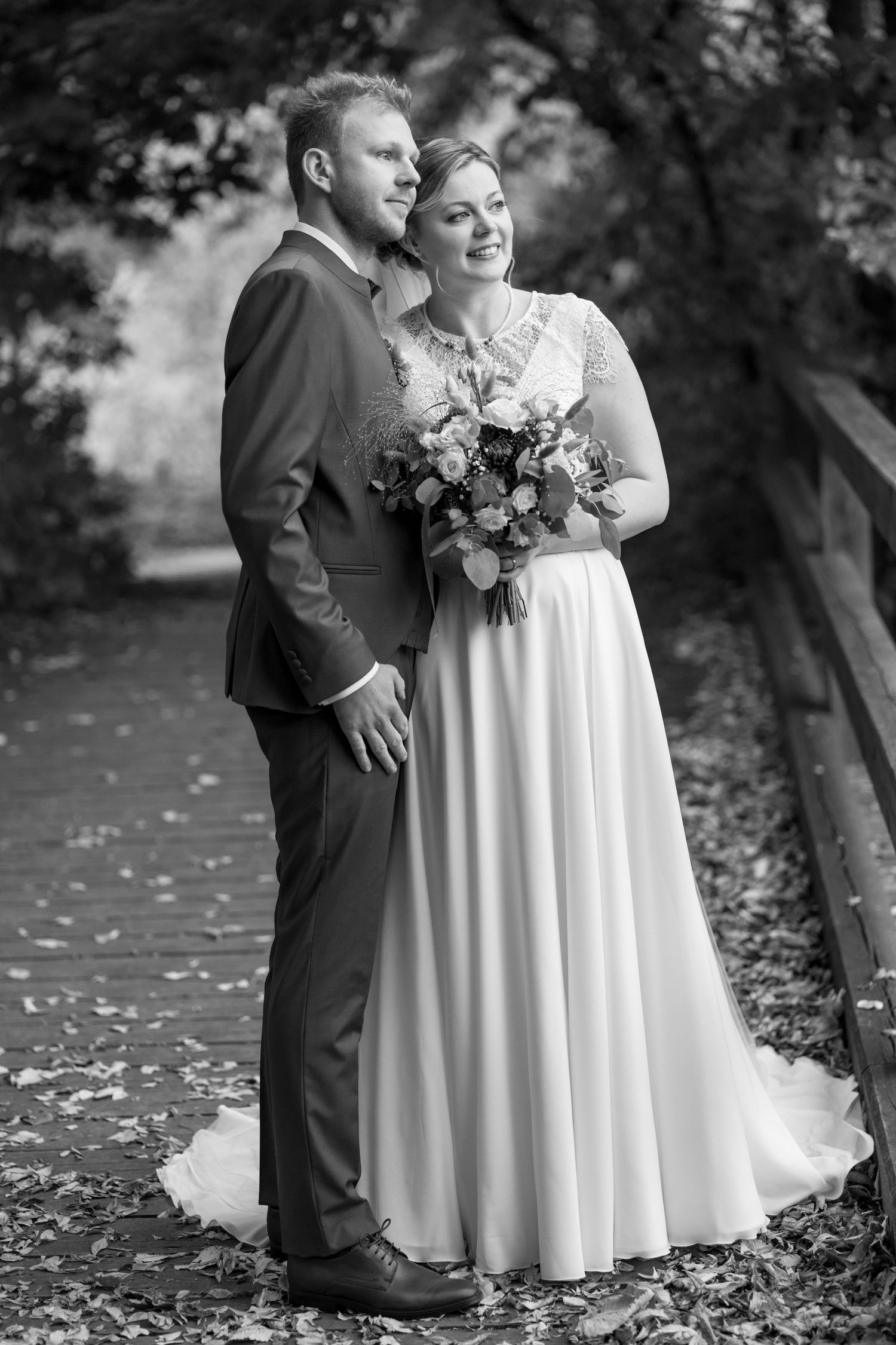 Black and white portrait of a couple in wedding attire standing on a leaf-covered path.
