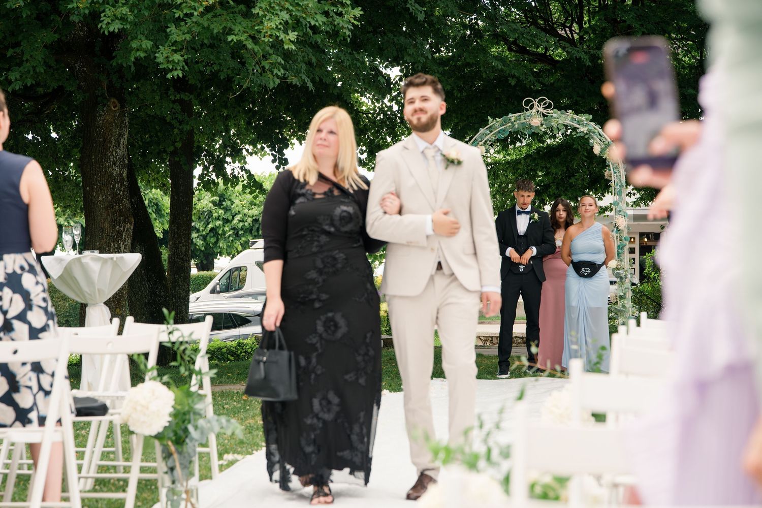 Groom in beige suit walks with woman in black dress at outdoor wedding ceremony with white chairs.
