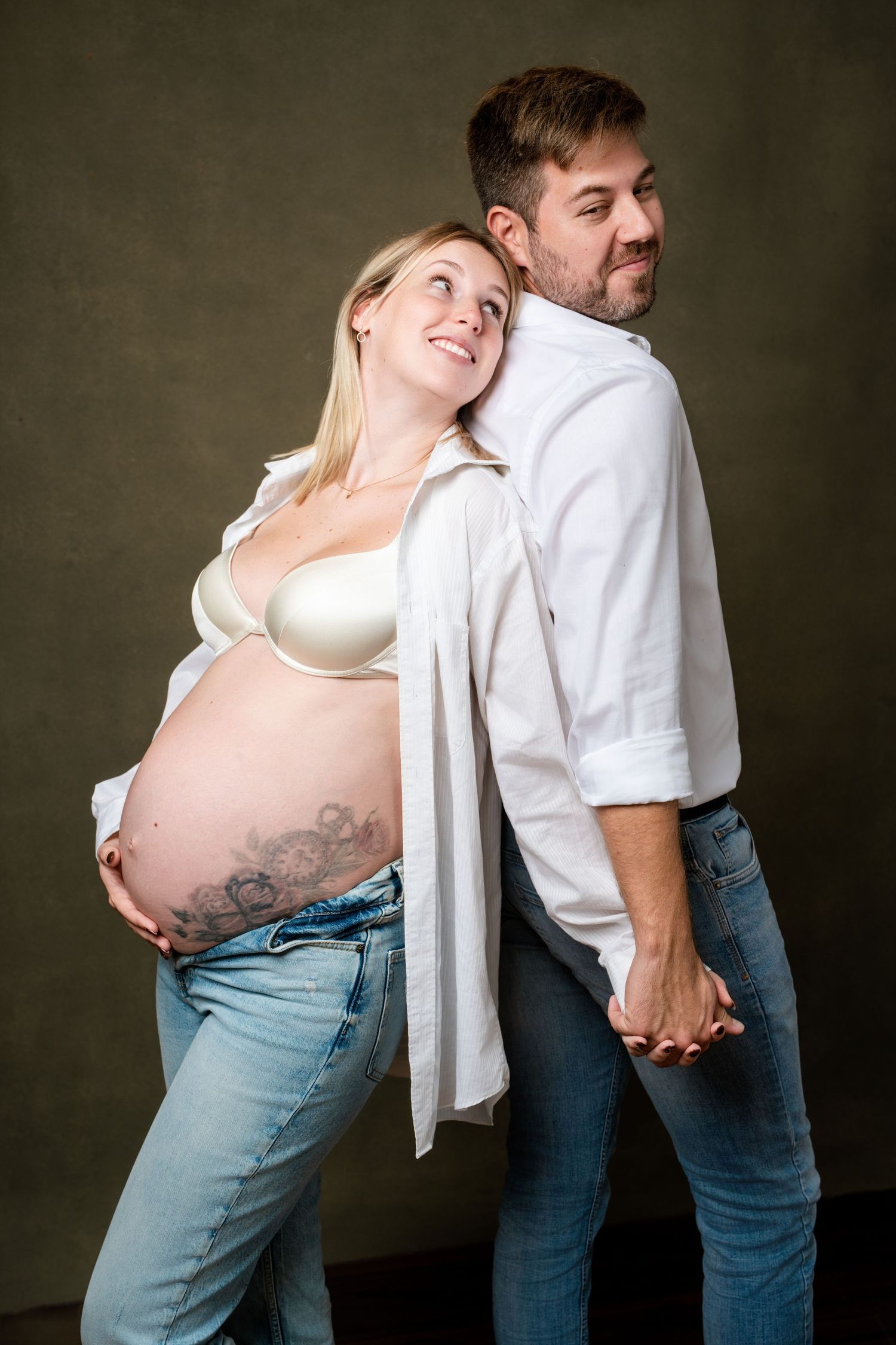 Intimate maternity photography showing couple in white shirts and denim against dark wall.