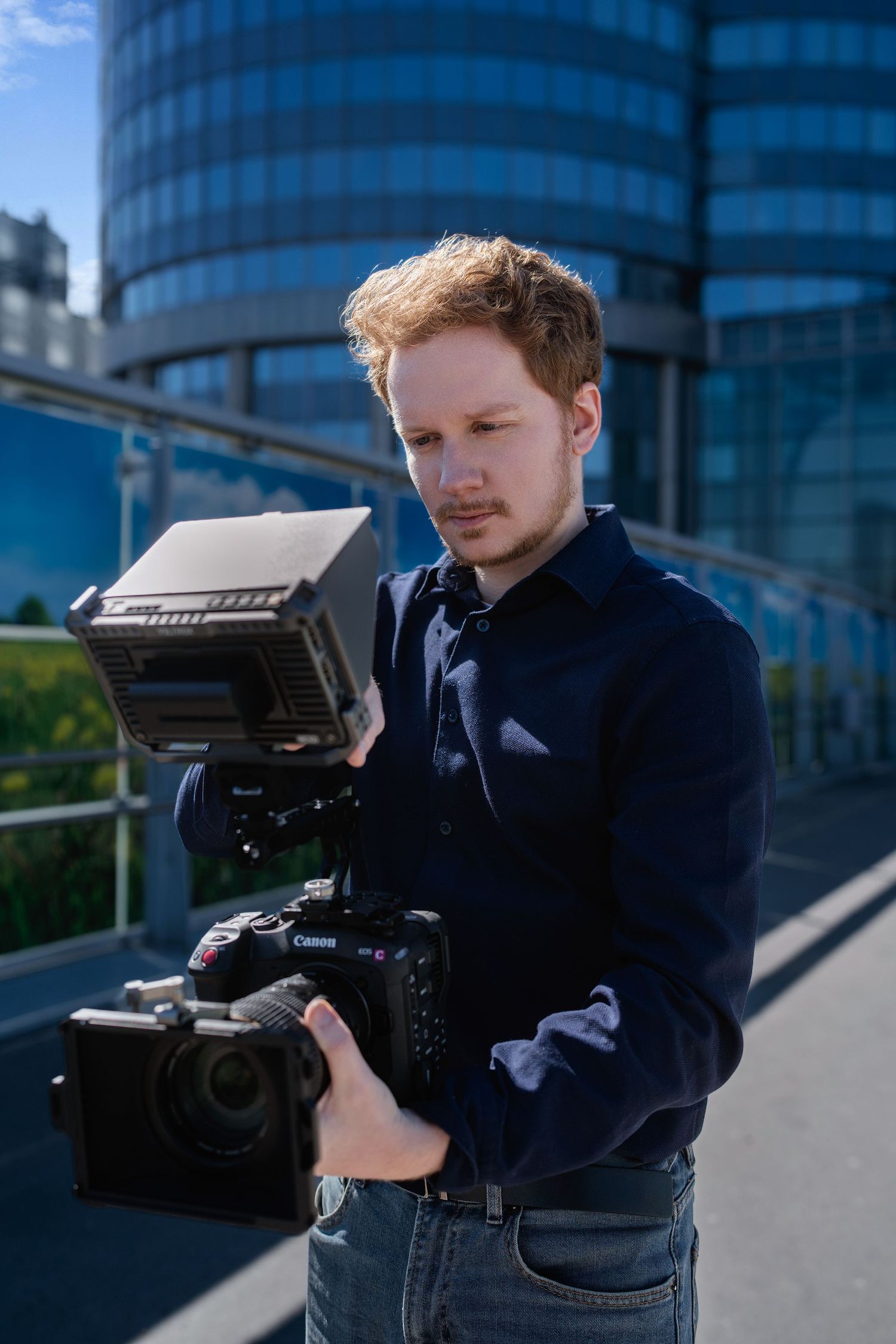 A camera operator wearing a navy sweater films with professional equipment against a modern urban backdrop.