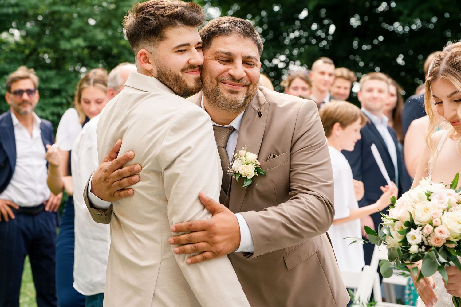 Two men in suits share a warm hug at an outdoor wedding ceremony with guests gathered in the background.