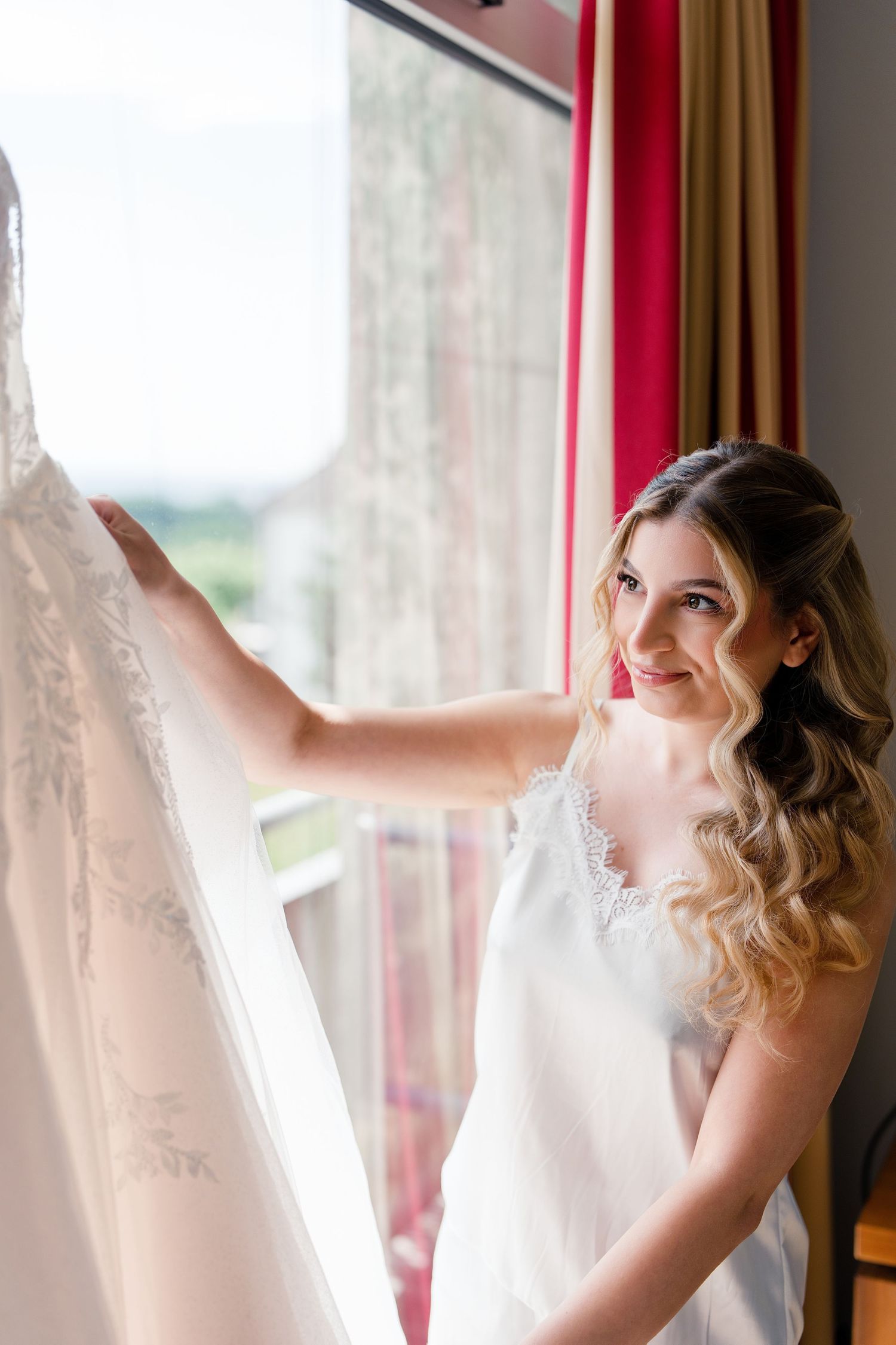 Bride in white slip dress smiling while touching her wedding gown hanging by a sunlit window.