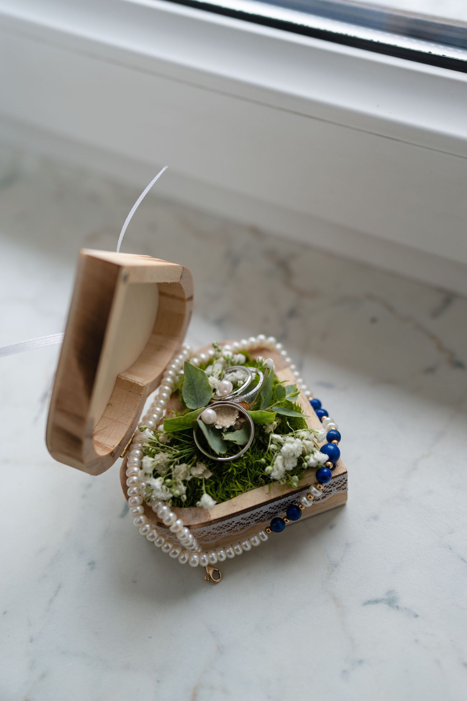 Wedding rings nestled in moss inside a decorative pearl-trimmed box on a white marble surface.
