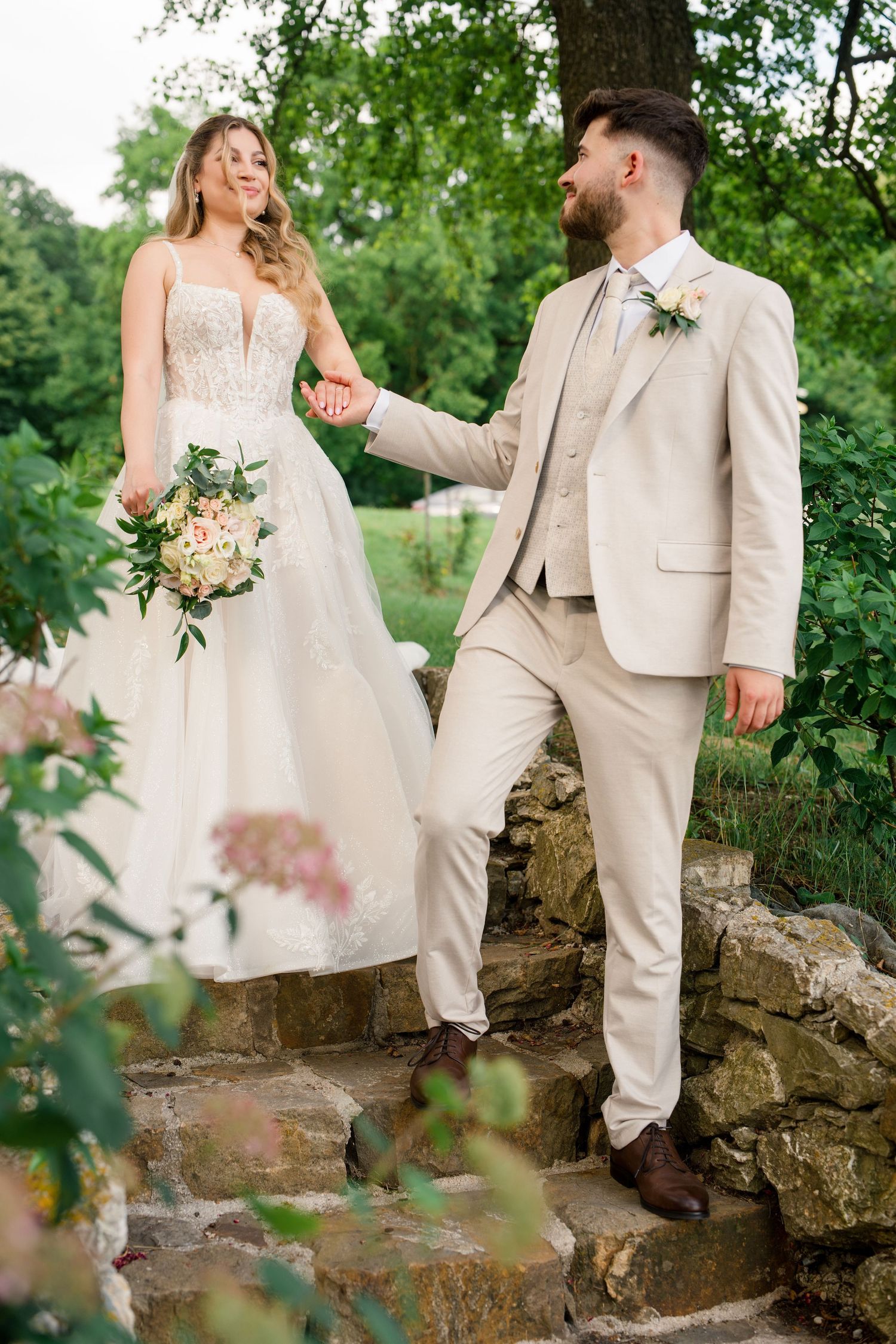 A smiling bride in a white gown holds a bouquet while a bearded groom in a beige suit holds her hand on stone steps outdoors.