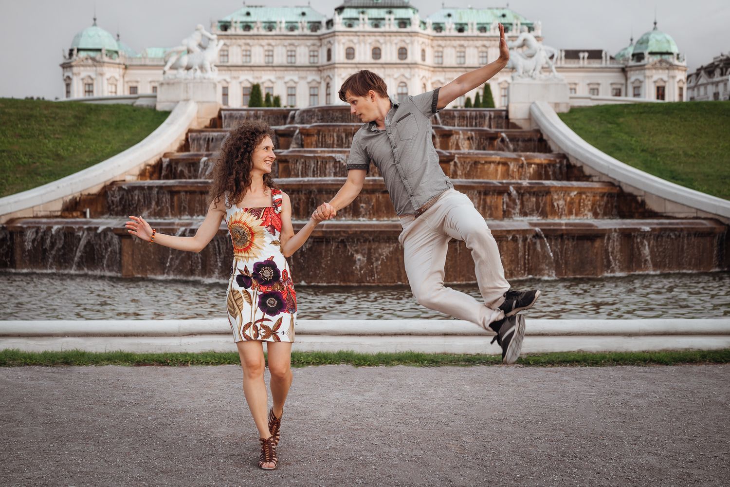 A romantic couple dances and poses near ornate fountains and architecture at a grand palace.