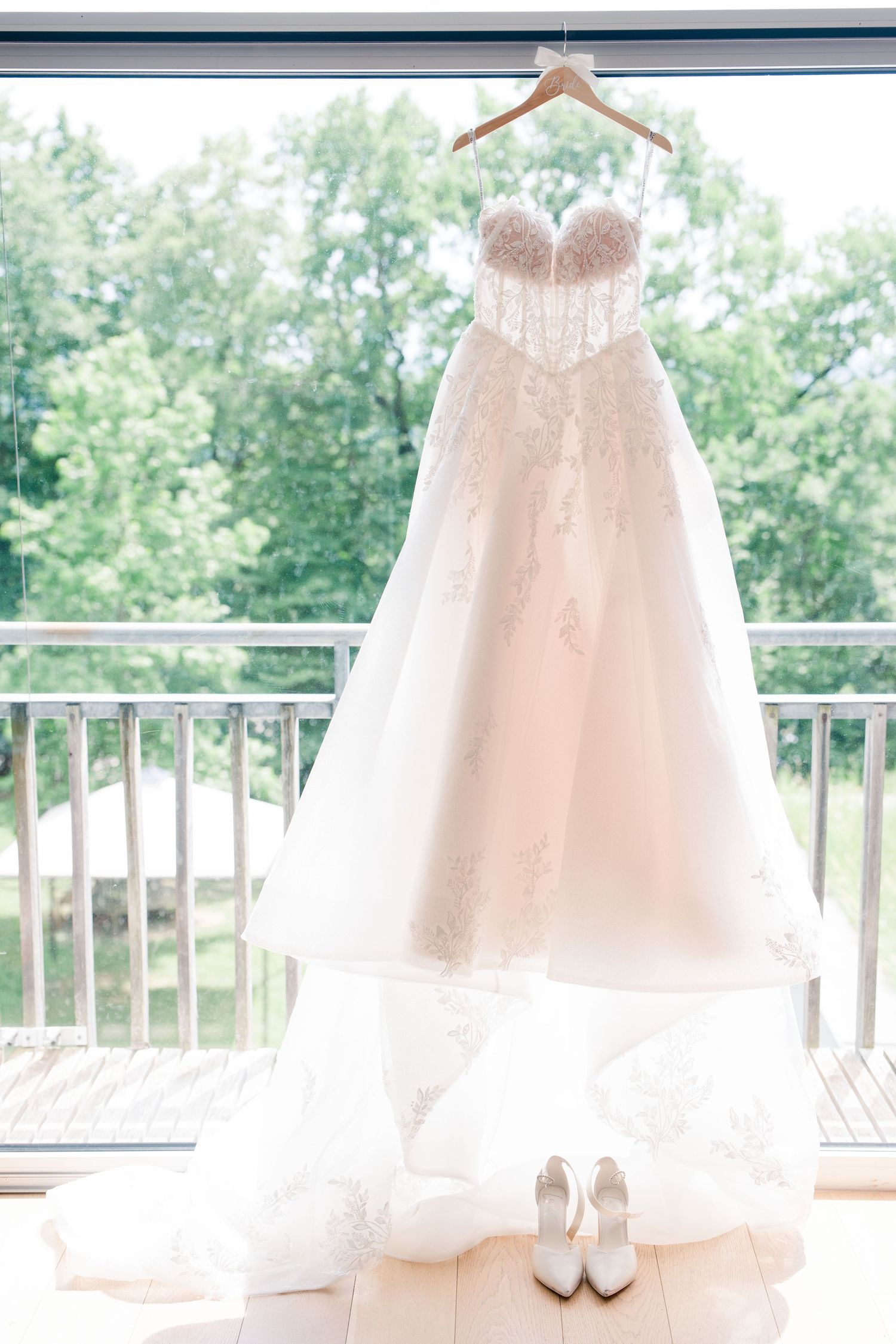 Elegant lace A-line wedding dress hanging on wooden hanger before window with white heels below.