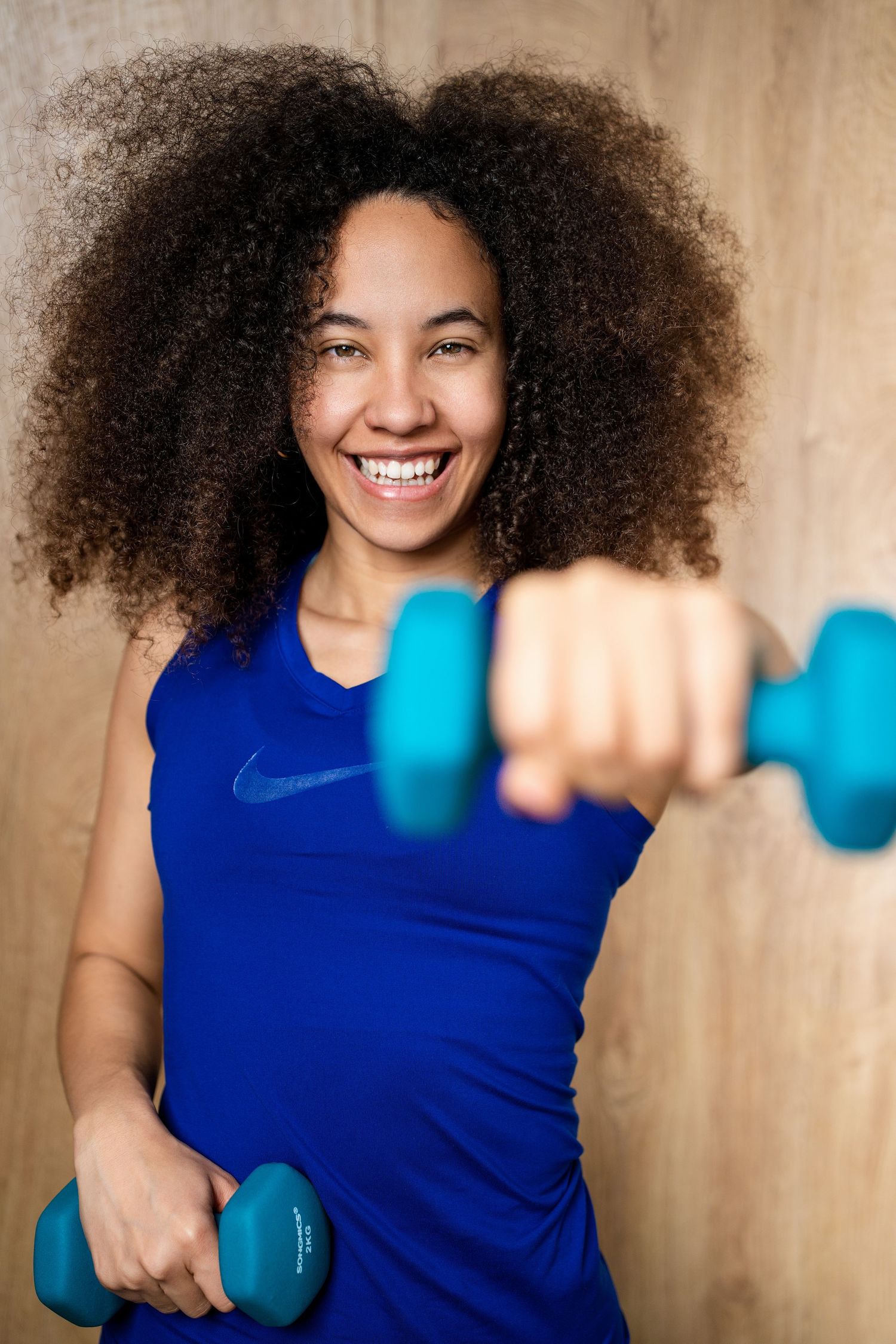 Smiling young woman with curly hair in blue Nike top holding teal dumbbells, extending one toward camera.