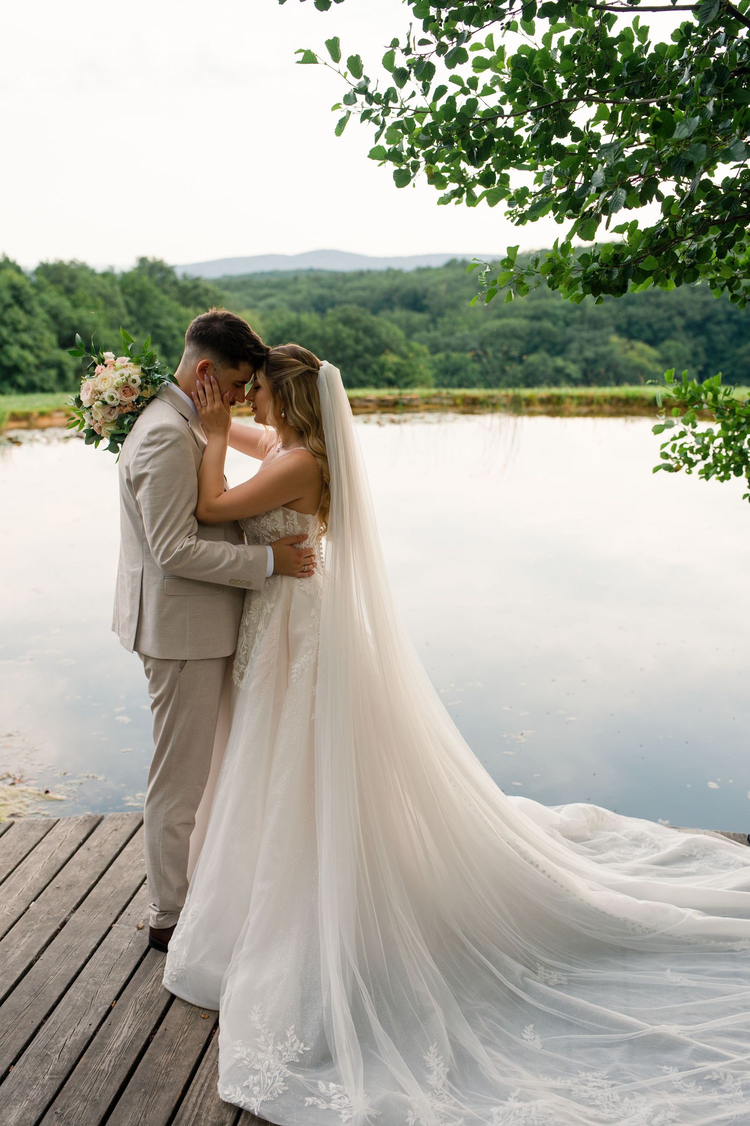 Bride and groom share a romantic embrace on a wooden dock by a serene lake, surrounded by lush green trees.