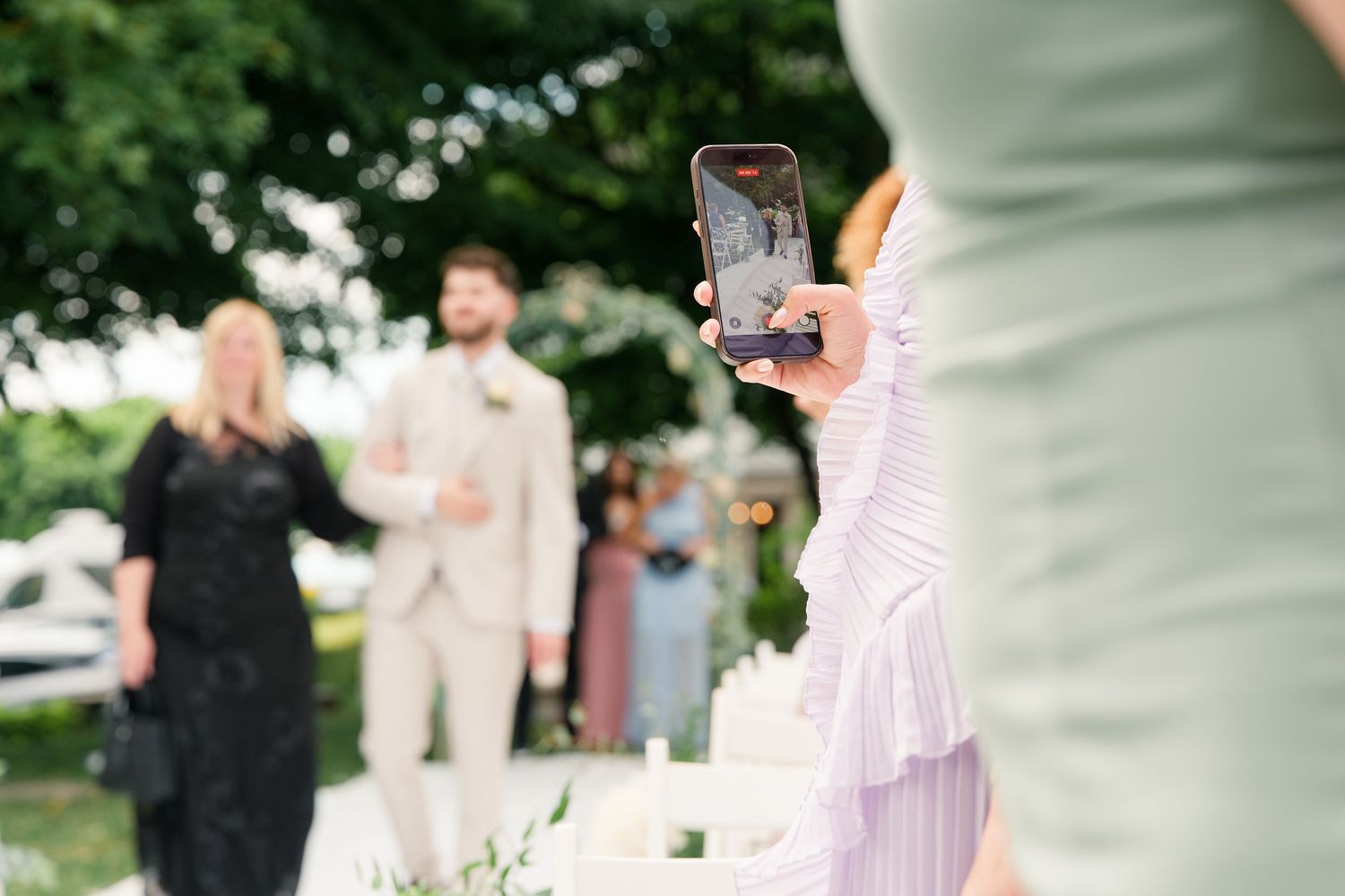 Guest capturing wedding ceremony on smartphone, with groom in tan suit and guests gathered outdoors.