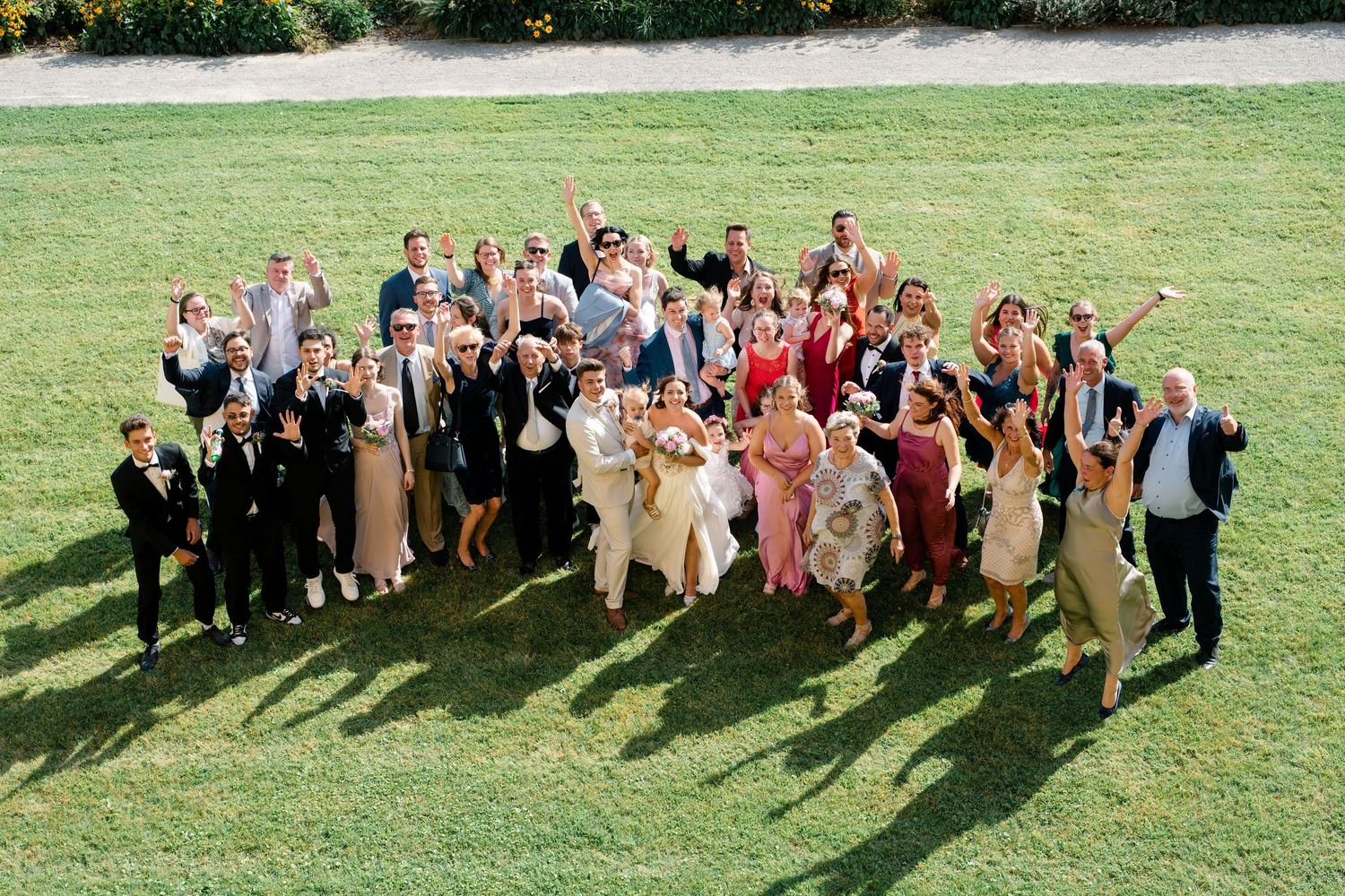 Large wedding party poses together on a sunny lawn creating long shadows.