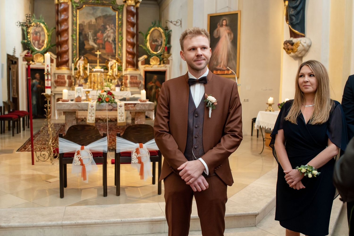 Two people stand at the altar of an ornate church, with one wearing a brown suit and the other in a black dress.