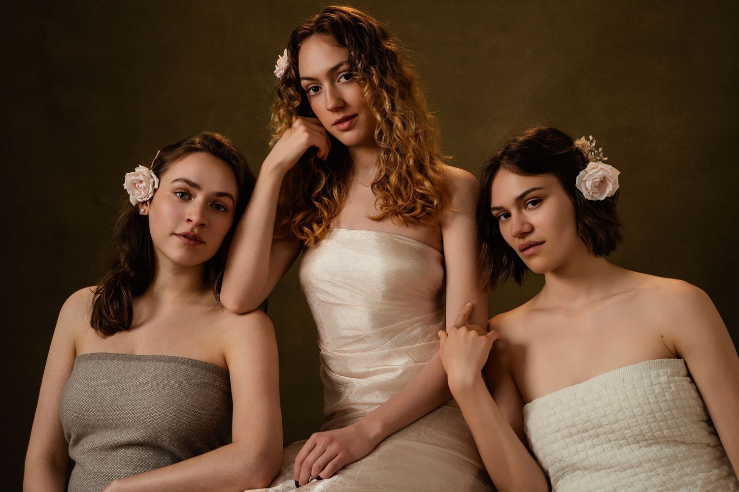 A group of women in elegant cream and beige dresses pose together in a moody, artistic studio portrait.