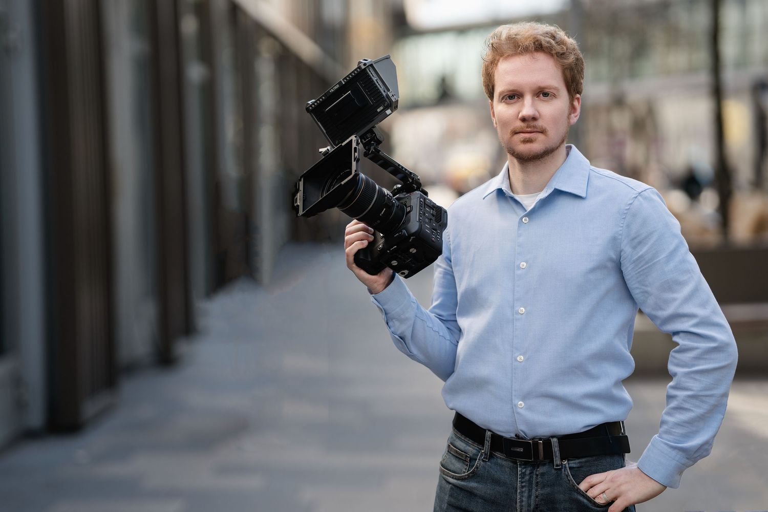 Professional photographer wearing light blue shirt stands with camera equipment in urban setting.