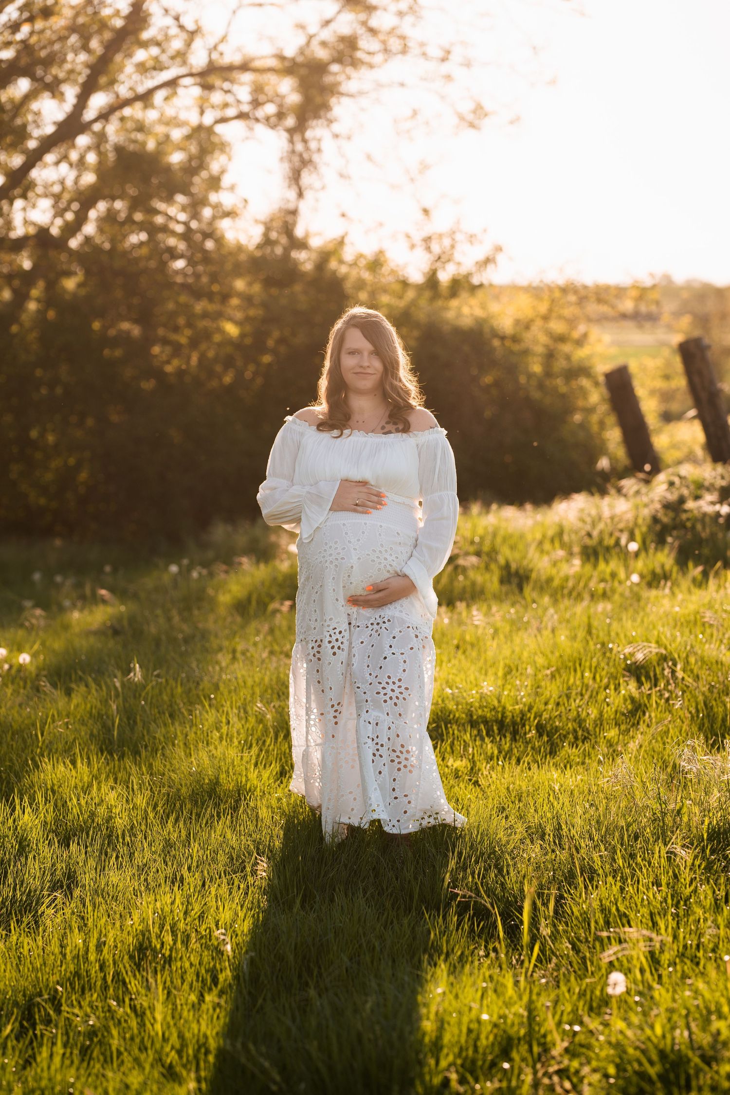 A person in a white lace dress stands in a sunlit meadow during golden hour, creating a dreamy silhouette.