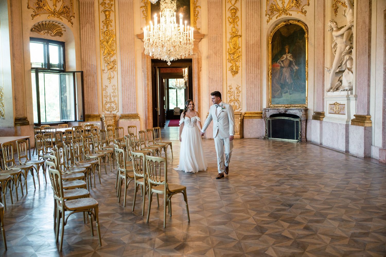 Wedding couple walks through ornate ballroom with crystal chandeliers and gold details.