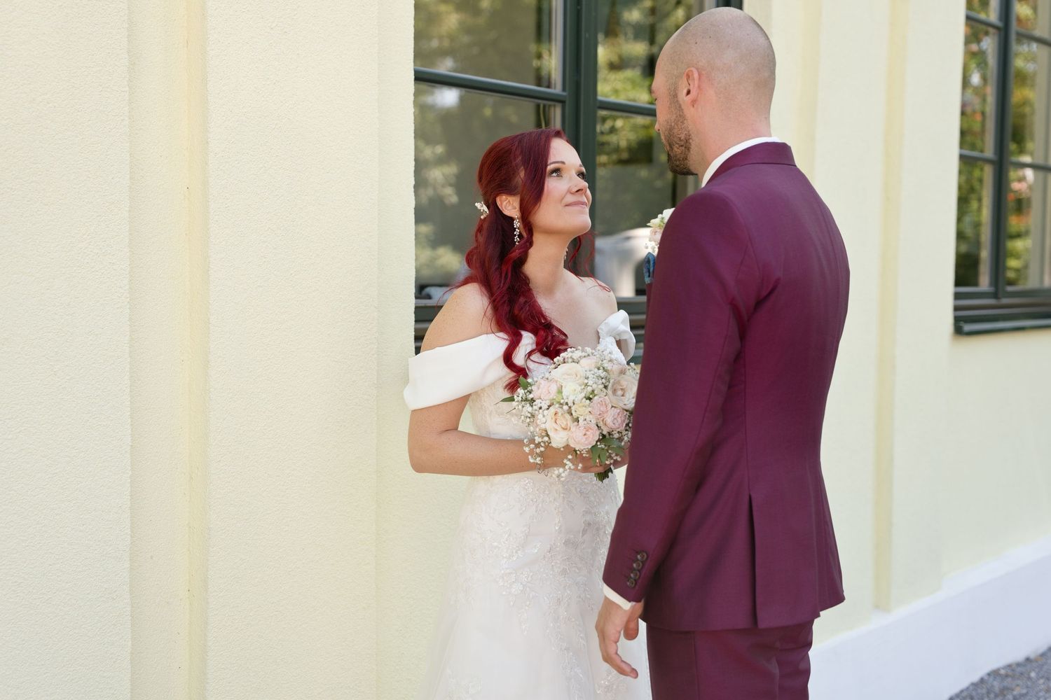 Bride in white off-shoulder gown holding bouquet smiles at bald groom in burgundy suit outdoors.