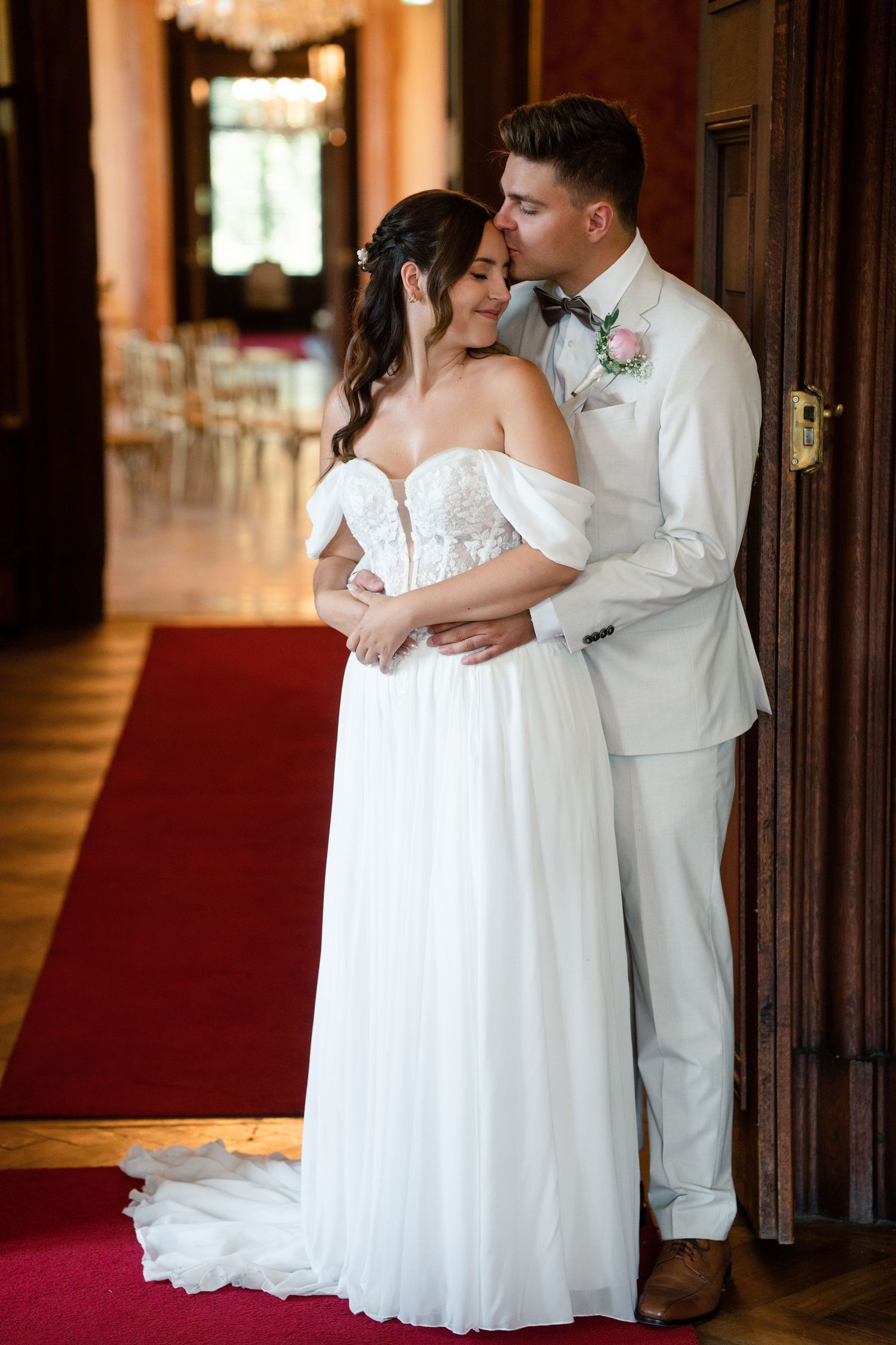 Elegant wedding couple embraces in hallway with red carpet and chandeliers.