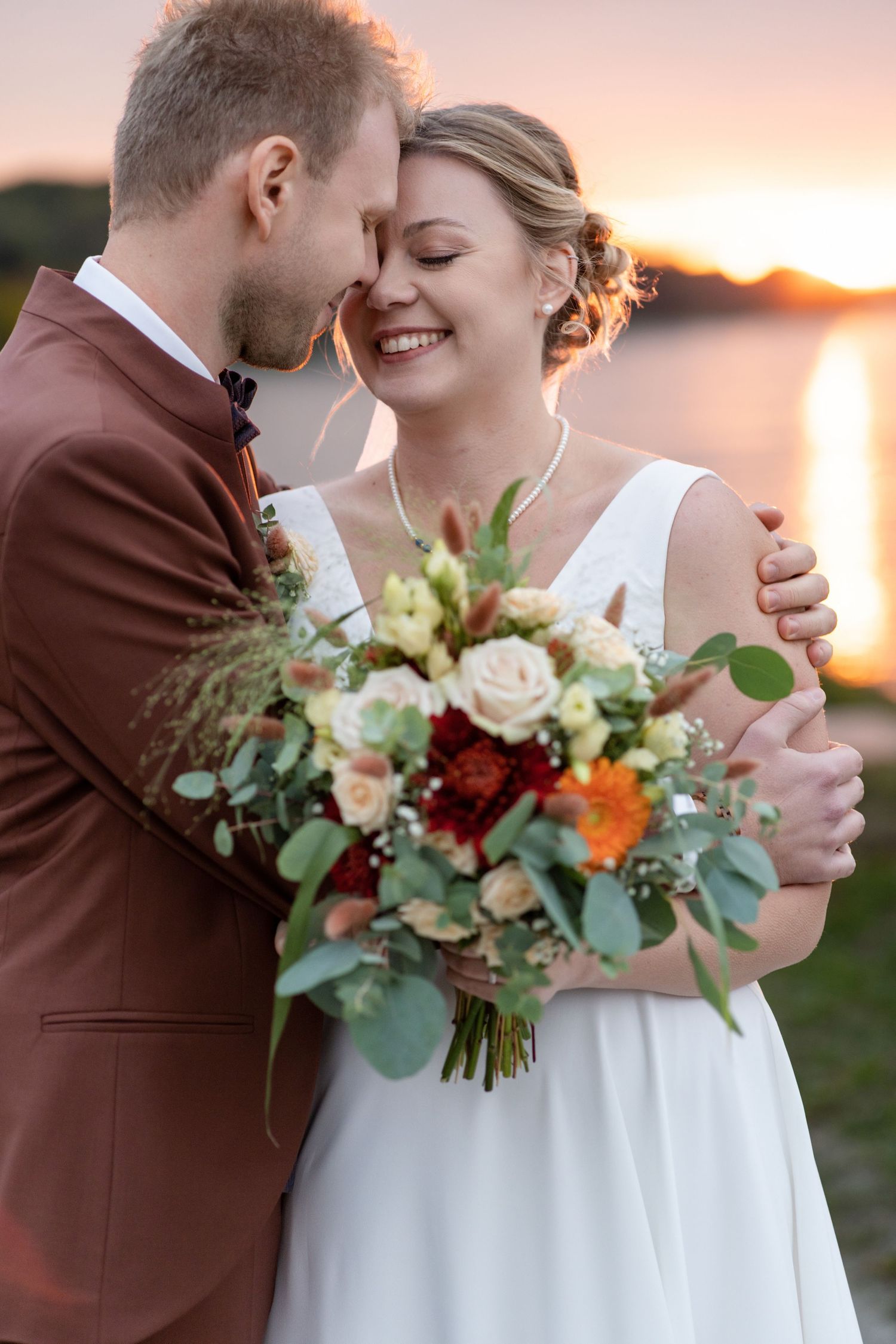 A romantic sunset wedding photoshoot featuring a burgundy suit and white dress with floral bouquet.