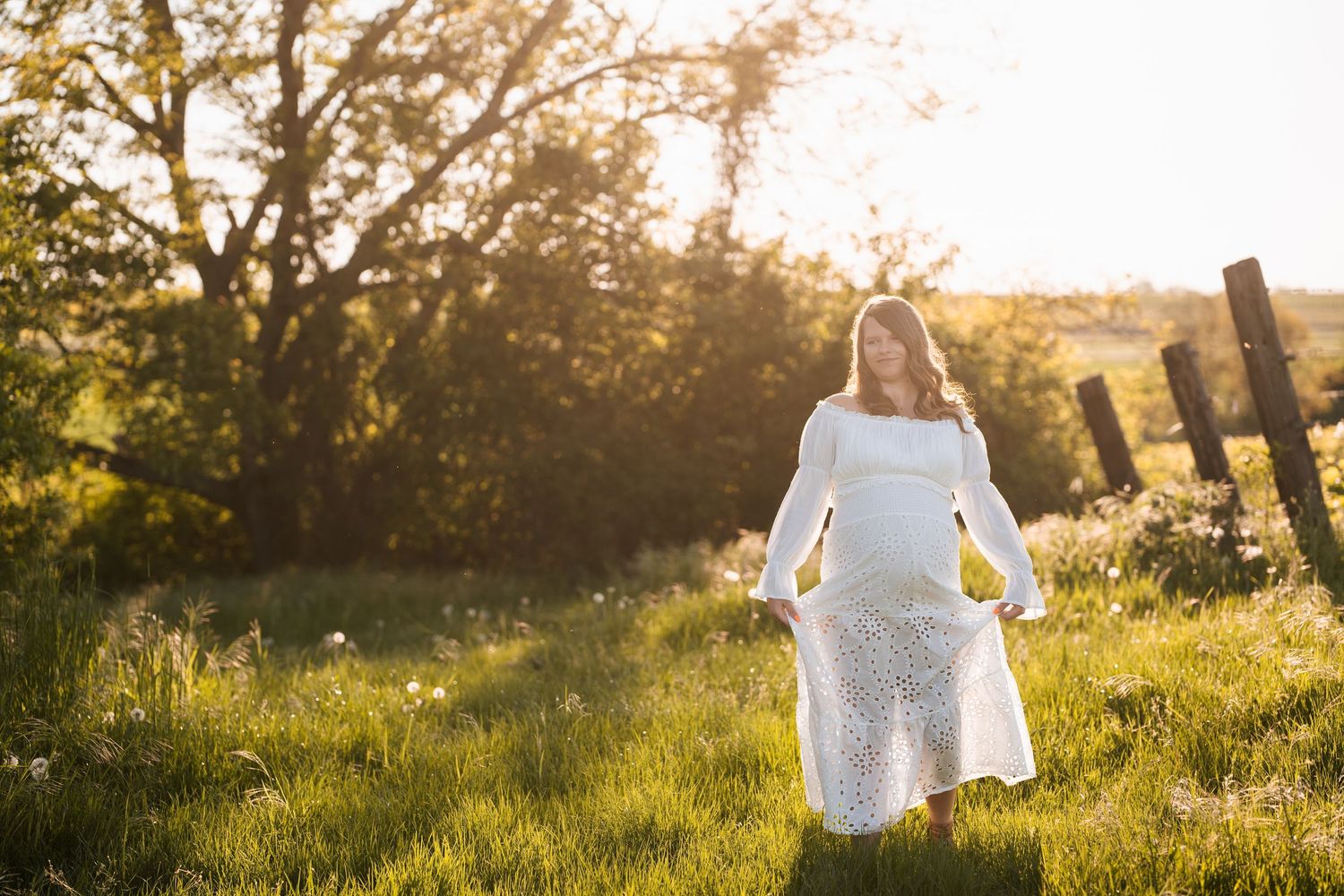 A person in a white dress walks through a sunlit meadow at golden hour with tall grass and wildflowers.